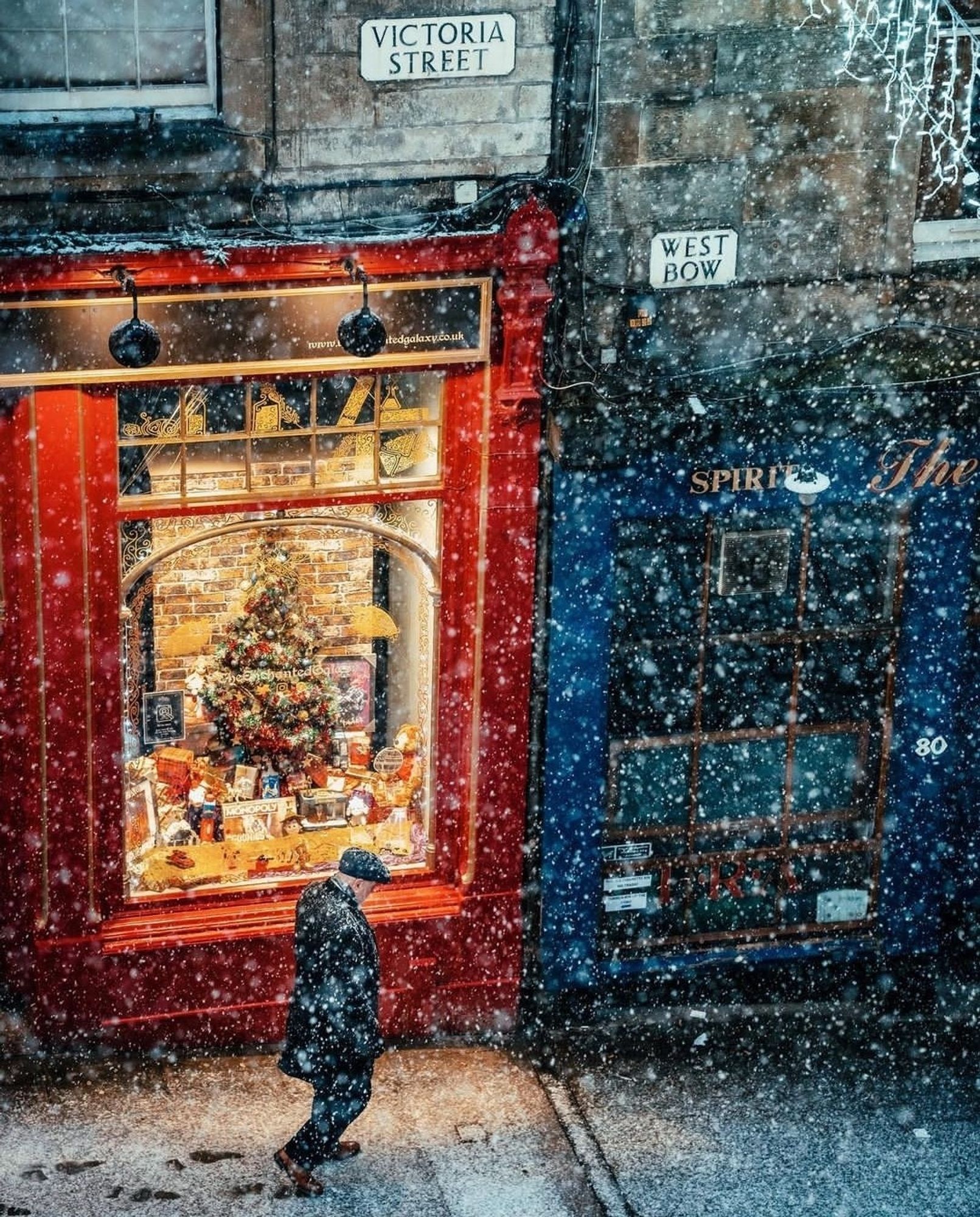 Photography. A color photograph of a shopping street in Edinburgh. A gentleman in a winter coat and cap walks along the sidewalk in the snow past a shop window decorated for Christmas. The red-painted building is a toy store. In the illuminated shop window is a Christmas tree with toys decorated around it. Together with the falling snow and the old houses, this creates a lovely pre-Christmas atmosphere.
Info: Victoria Street is one of Edinburgh’s most recognisable and picturesque locations, its elegant curve graced by a series of colourful shop fronts, making it a favourite choice of filmmakers and photograph enthusiasts alike. It reminds many Harry Potter fans very much of Diagon Alley.