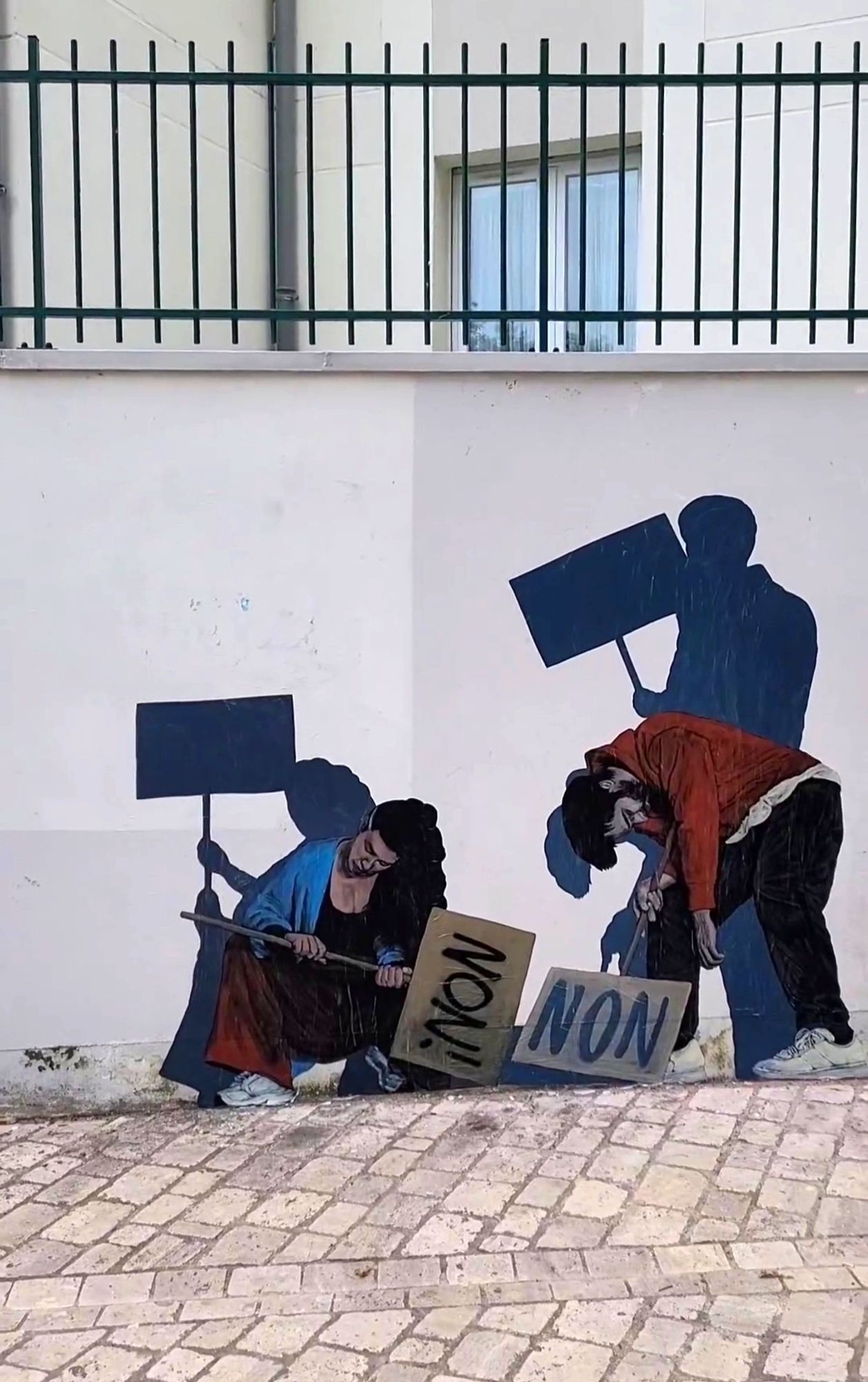 Streetartwall.  A self-painted pasteup of two sleeping people is stuck on a white wall with bars above it. A dark-haired man with a full beard is standing with a protest sign next to a sleeping dark-haired young woman. He is wearing red trousers, a black sweater and sneakers. She is sitting on the floor wearing red trousers, a blue vest and a black T-shirt. Both are holding signs with the French word "non" (English: no) written on them. Behind them are their shadows. They are not sleeping and are holding up the signs.
Info: Notes by the artist Levalet: "In these troubled times, we can remind ourselves that each of us has the strength to say no, to resist, even when the worst seems certain. Let's wake up!"