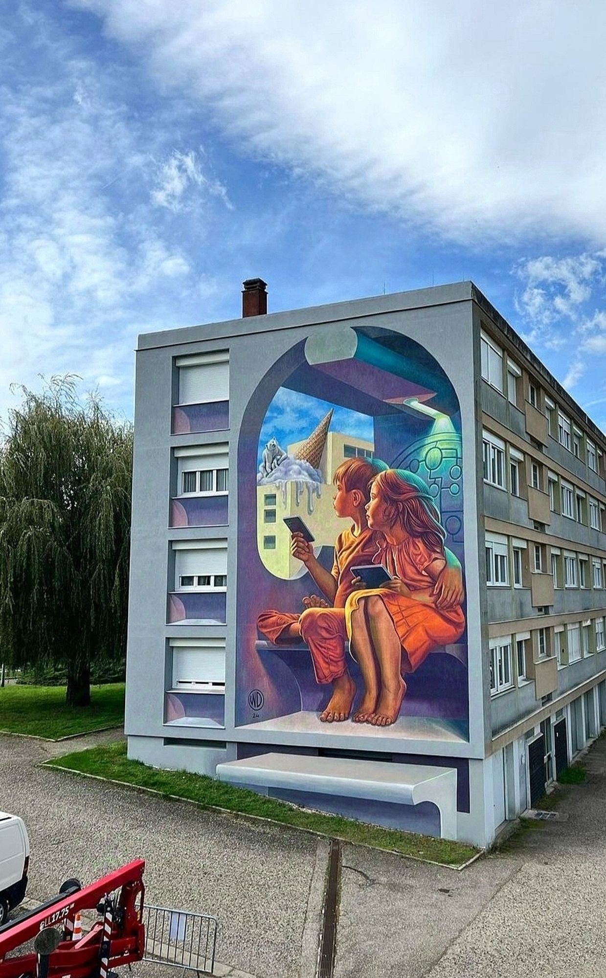 Streetartwall. The image shows a large, imaginative mural on the side of a long, four-story building. The mural shows two children (a girl and a boy dressed in orange) sitting close together in a tiny room, each holding a tablet in their hands. They are looking at a surreal scene in the background in which a polar bear is sitting on a melting ice cream cone. The mural is painted in 3D with abstract shapes and patterns. The room seems to tower over the wall. The mural is colorful and detailed, combining realistic and fantastical elements. (The artist's initials "WD" and the year "24" can be seen at the bottom left of the mural).
Info: The Artist about the Mural:
"While just 100 companies are responsible for over 70% of world's greenhouse gas emissions - which cause global warming and lead to climate change - they still try to convince us that all is about individual responsibility."