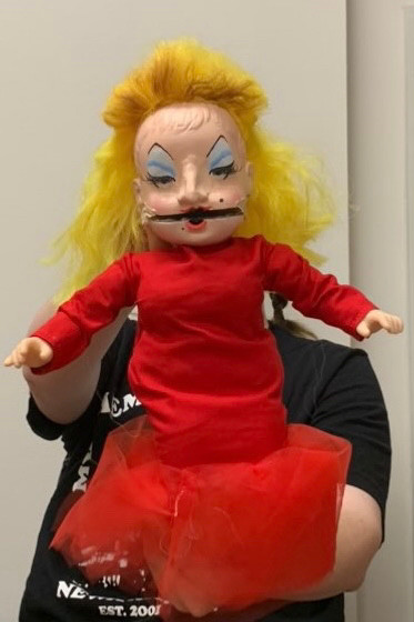 Puppet of Divine the drag queen in her red dress from Pink Flamingos. 

The puppet is made of a baby doll that's head has been cut in half. It has a bright yellow mullet and a form fitting red dress with tulle at the bottom. 

The person holding it up is almost visible, obscured by the puppet. 