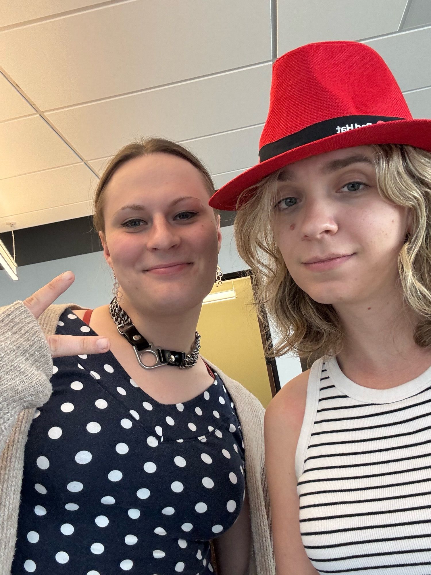 two women, one in a polkadot top with a suspicious chain collar, the other in a striped tank top and a bright red RedHat branded fedora