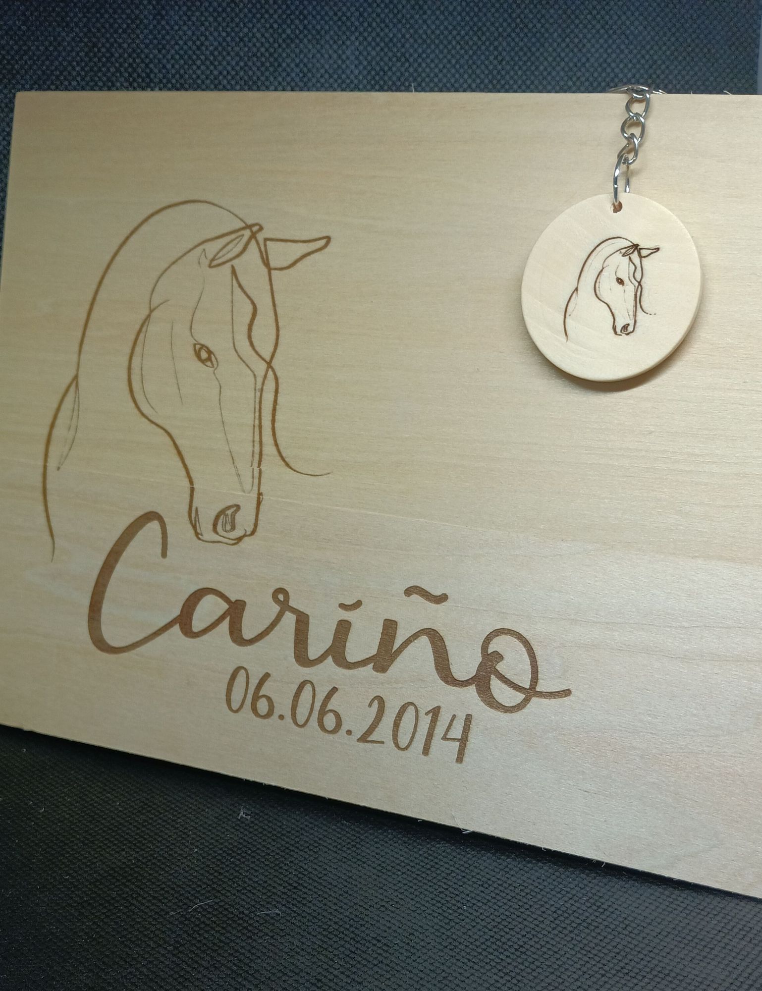 Ein Holzschild mit einer Einlinienzeichnung eines Pferdekopfes. Darunter steht in verschlungener Schrift Cariño und das Datum 06.06. 2014.
In der rechten oberen Ecke hängt ein kleiner Schlüsselanhänger mit demselben Bild.