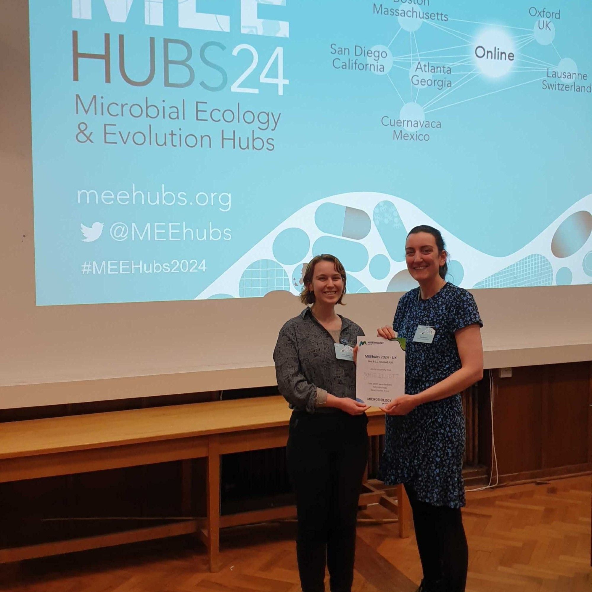 two women holding an award certificate, standing in front of a conference presentation slide