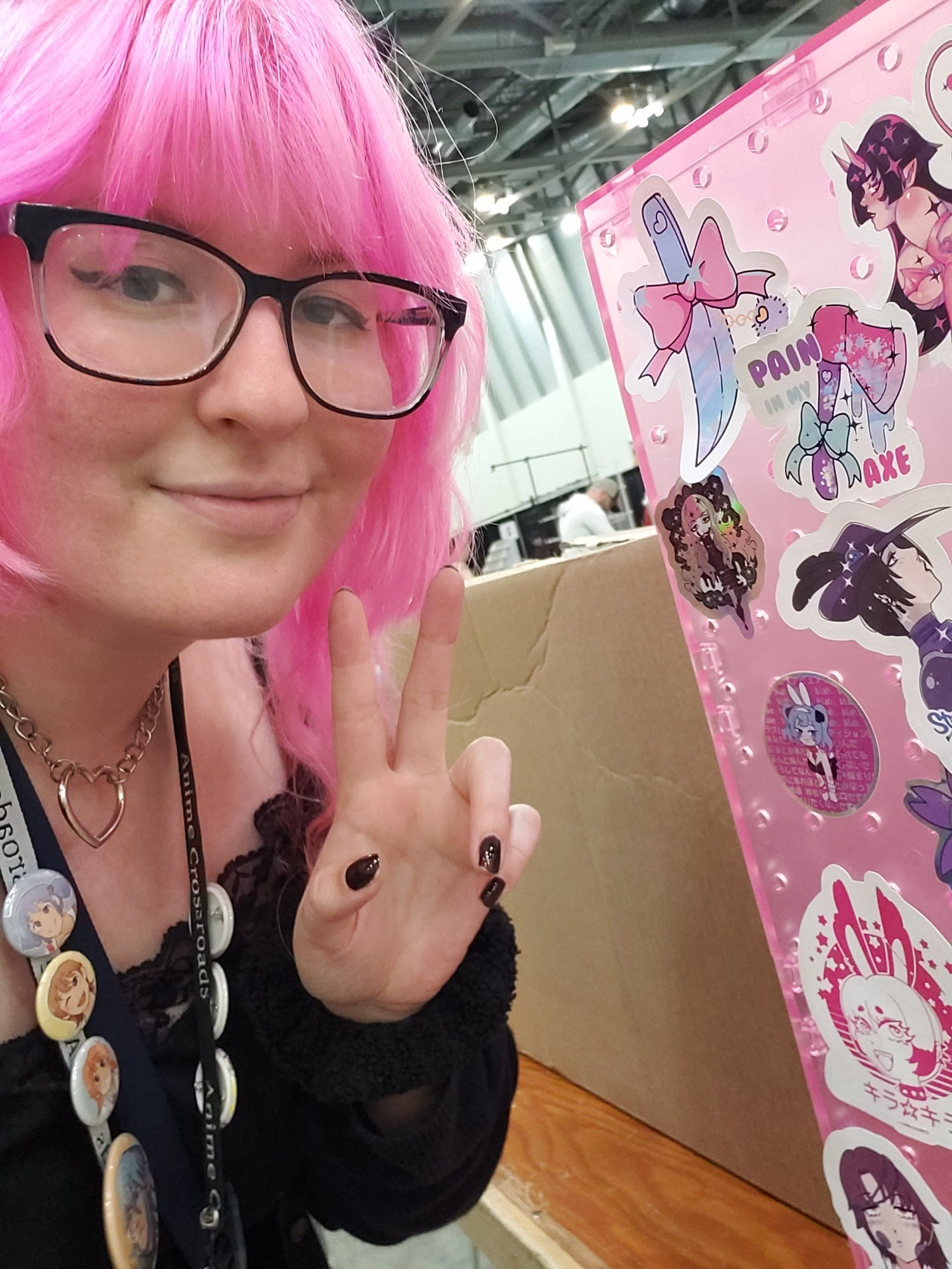 Selfie of candlehearts, a pink haired femme making a peace sign with their hand. On the side is a pink pegboard with stickers and a box behind