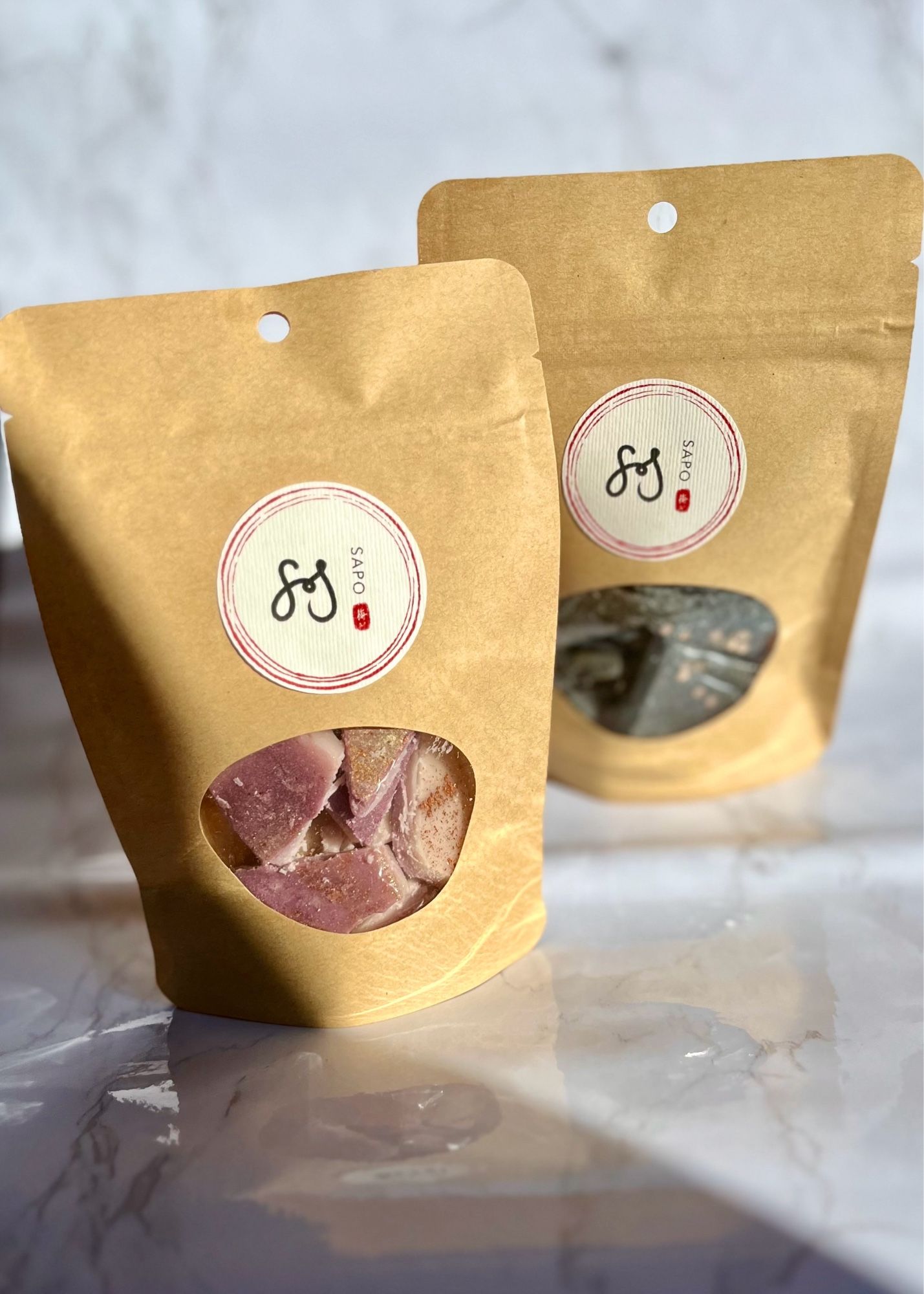 two commercially compostable kraft bags of wax melt brittle in the scents honey roasted figs and cemetery stroll sit on a pale faux marble backdrop in the sunlight