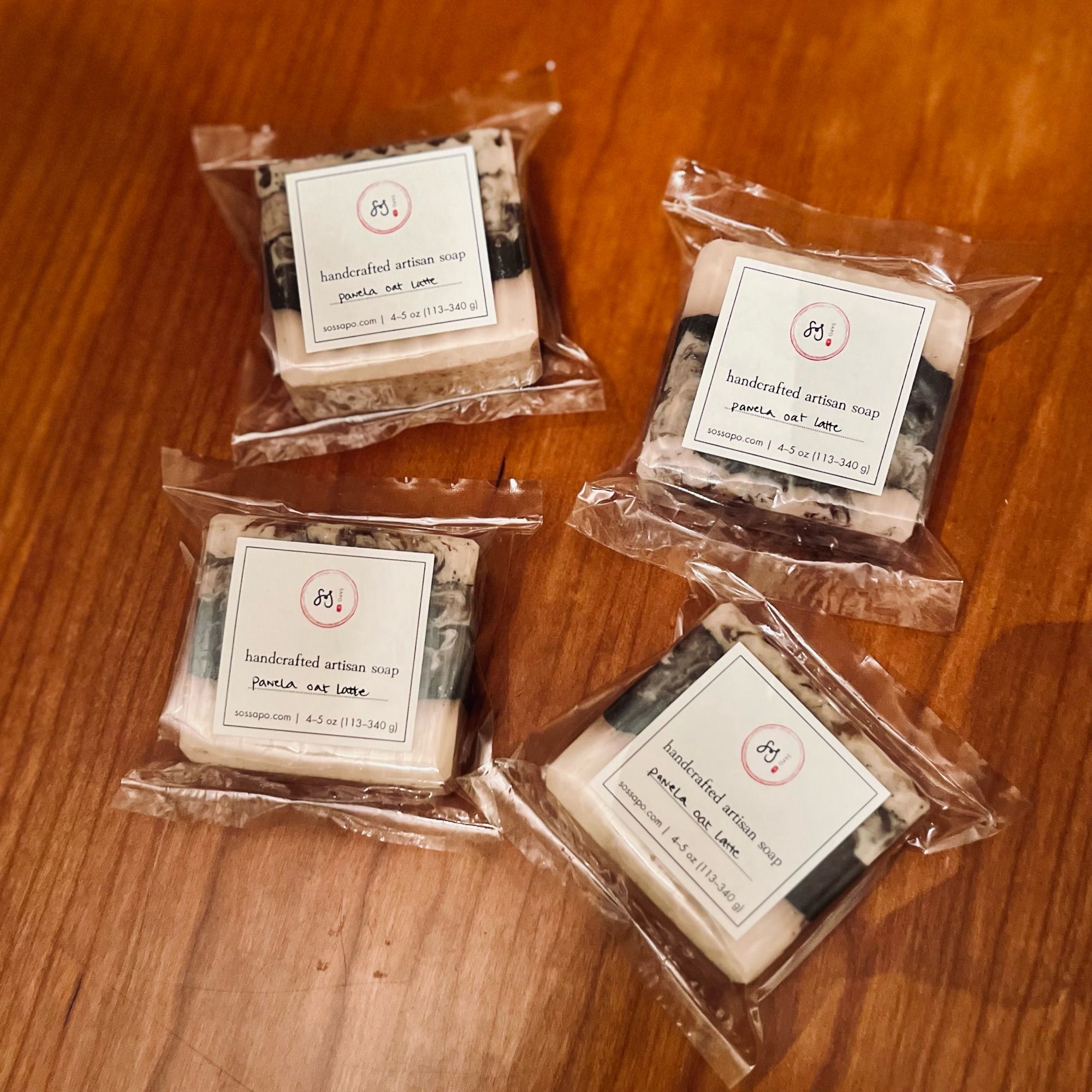 four 4 oz panela oat latte soaps in home compostable cellophane bags sit atop a cherry wood table