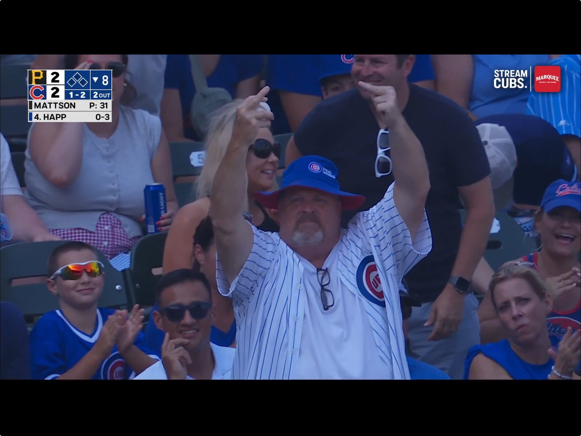 A goateed Cubs fan in the stands at Wrigley field wearing a jersey and blue bucket hat, emphatically flipping off the camera with both hands