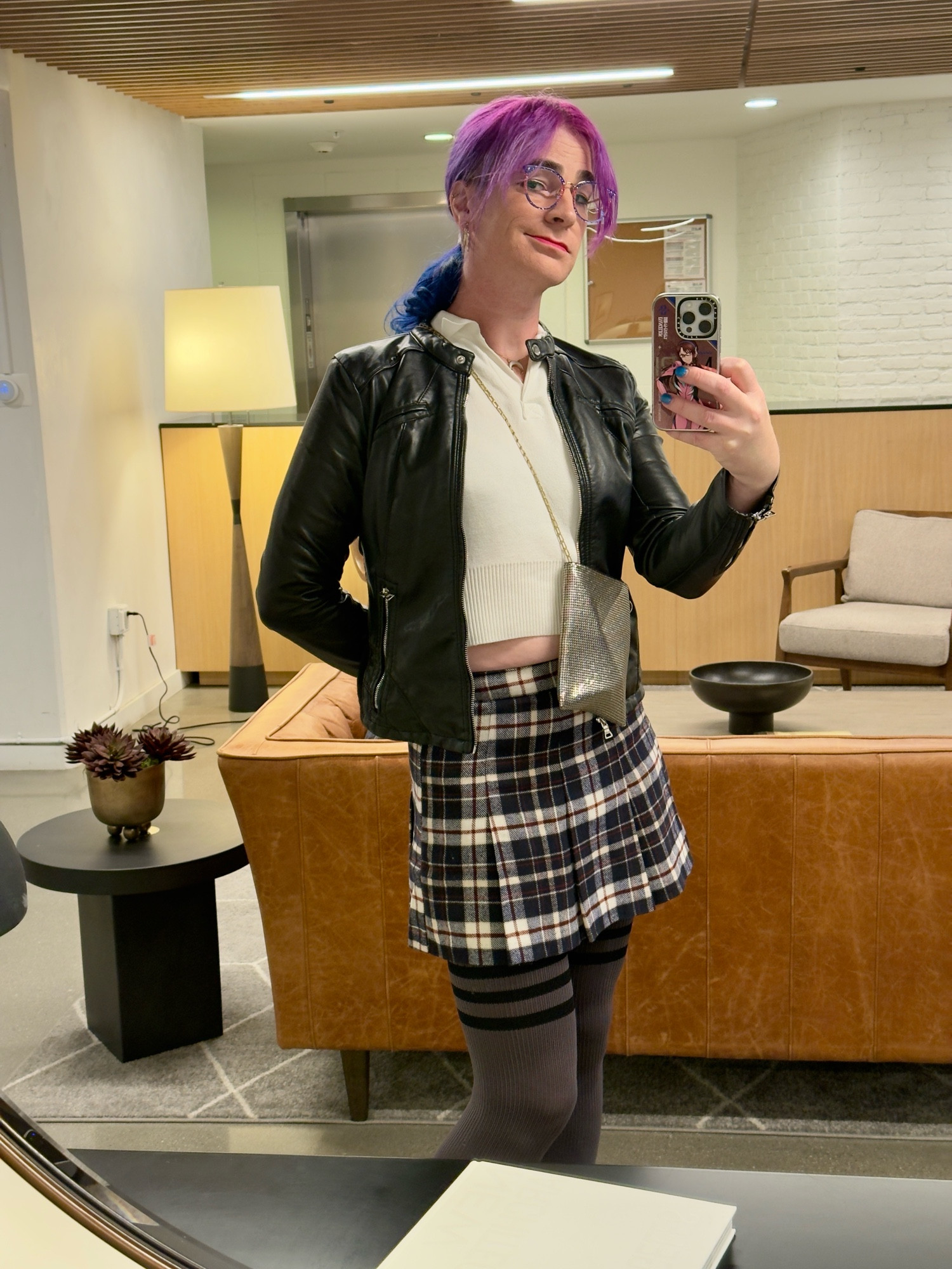 becs, standing in a lobby & taking a selfie in the mirror. she's hot pink and blue hair tied into a ponytail, and wearing a blue and white skirt with a white cropped polo under a black leather moto jacket