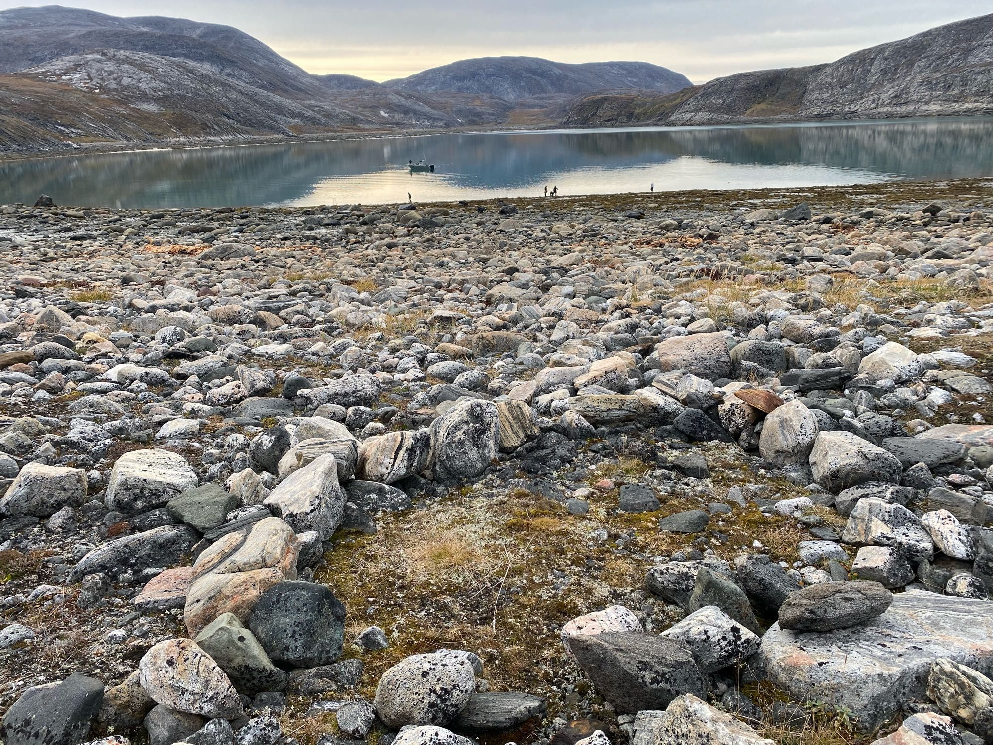 A mountain surrounded bay with a rocky foreground.  A cleared circle exposing tundra was a campsite— perhaps ancient?  Figures on the beach are gathering mussels.