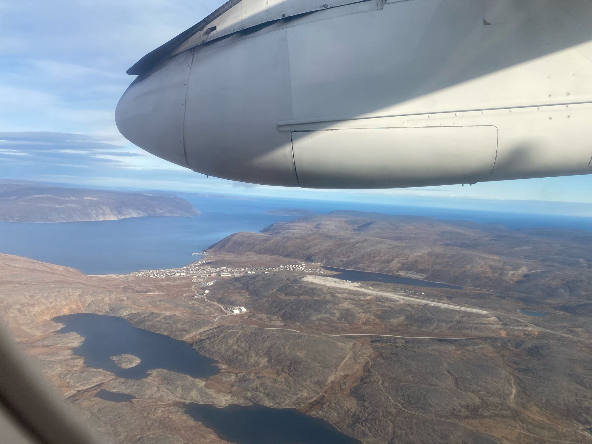 Aerial view of the town of Kangiqsujuaq, with Wakeham Bay in background.  Lakes and rocky tundra in foreground.  The gravel airstrip is right below the Dash 8’s engine nacelle.