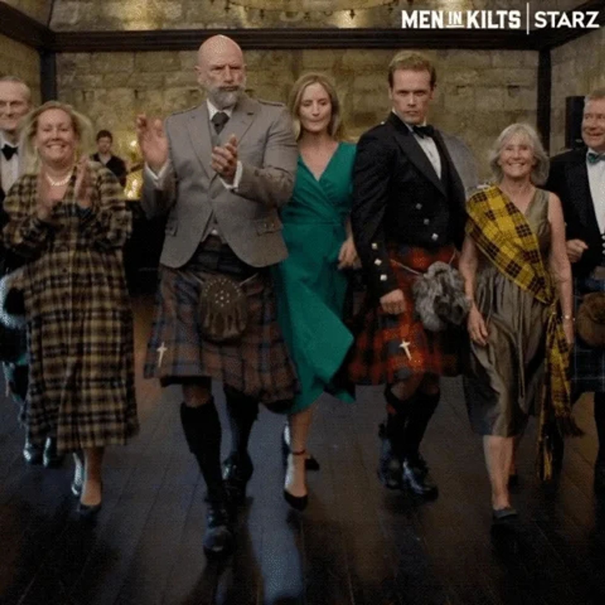 People joyful dancing, starting on the left: a blonde woman in a long yellow black tartan dress which is long / covers knees. Graham McTavish in a grey jacket, brown / blue tartan kilt, a kilt sporan, knife pin and black socks and shoes. A blonde woman in a green dress. Sam Heughan in a black jacket, with red tartan kilt, a kilt sporran and black socks and black shoes. And a woman in a olive green dress with a yellow tartan scarf across her chest.