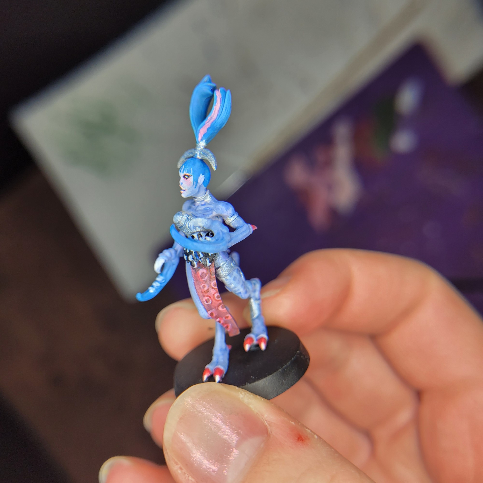 A slaanesh warhammer minifigure held up to the camera in Theo's hand. This minifigure has light blue skin but otherwise takes heavy inspiration from the musical artist Ashnikko. Her hair is in a metallic silver ponytail, with the hair arching upwards in two bright blue ponies that are reminicent of Horns. She has a black and white checkered chest piece with a silver metal breast cup, as well as a loin cloth painted to look like an octopus tentacle. Her foot claws are hot pink and her crab like hand claws are short and fade from her light blue skintone into the same bright blue as her hair.