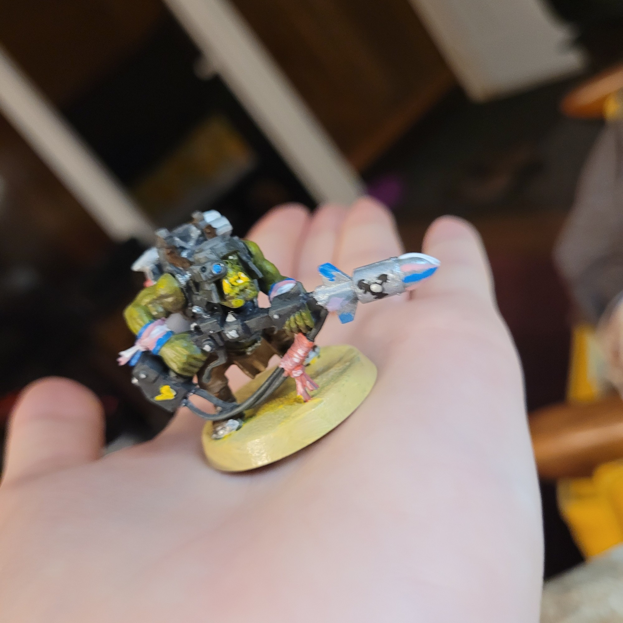 A slightly blurry picture of a painted ork warhammer minifigure resting on the palm of Theo's hand. The ork is painted in neutral greens and browns except for his arm bands and the missile at the end of his gun which are branded with the  olours of the trans flag, blue, pink, and white. The missile also has a small messily painted trans symbol on its side.