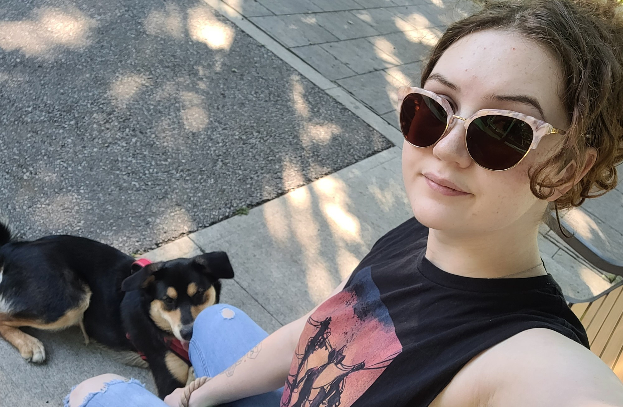 A selfie of Theo and their dog Lucky. In the photo Theo is sitting on a park bench with a small smile and sunglasses on. In one hand Theo holds the phone from a high angle and in the other hand holds Lucky's leash. Lucky is seen laying down comfortably on the sidewalk in a red harness. The two are under the shade of a tree with the sun peeking through the leaves casting golden pockets of light on the groud.