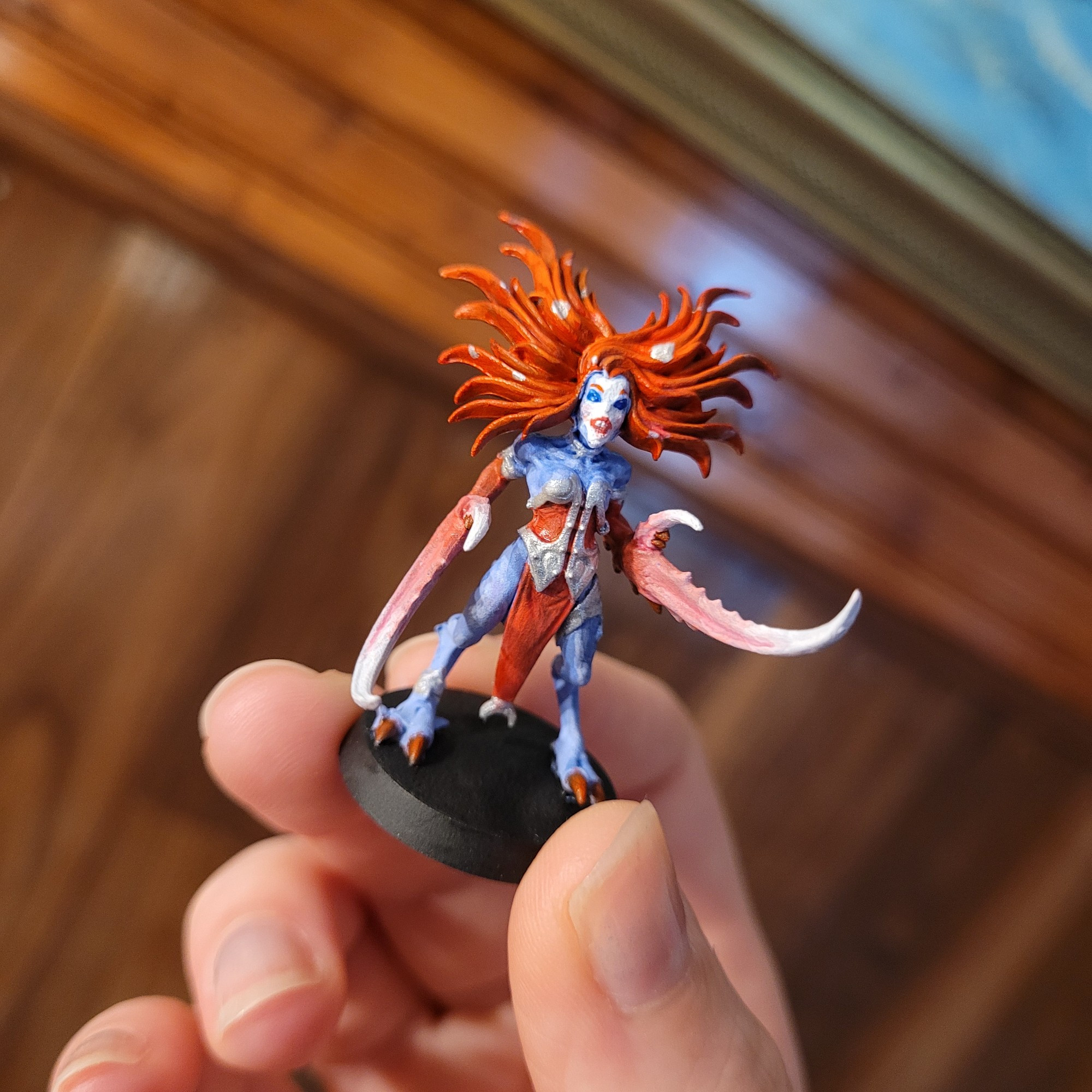 A slaanesh warhammer figurine held up to the camera in Theo's hand. The minifigure has the classic blueish slaanesh skintone but that is where the colour similiarities end. The minifigures hair fans out in a bright vibrant red, her armor is hot pink with silver metal details, her toenails, or rather toe claws are  hot pink as well. Her large crab like claws that take place where her hands should be are hot pink towards the elbow and fade into a pure white at the tips of her front claw and thumb claw.