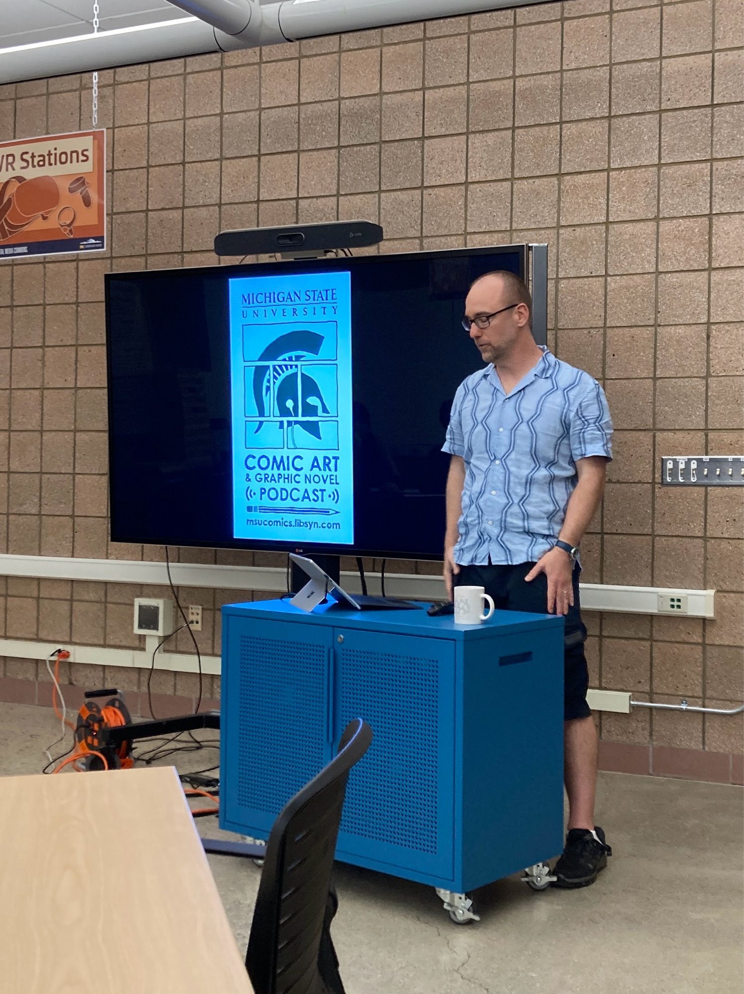 Ryan Claytor, standing at a low blue podium. Behind him on a large television screen is a slide advertising the MSU Comic Art & Graphic Novel Podcast