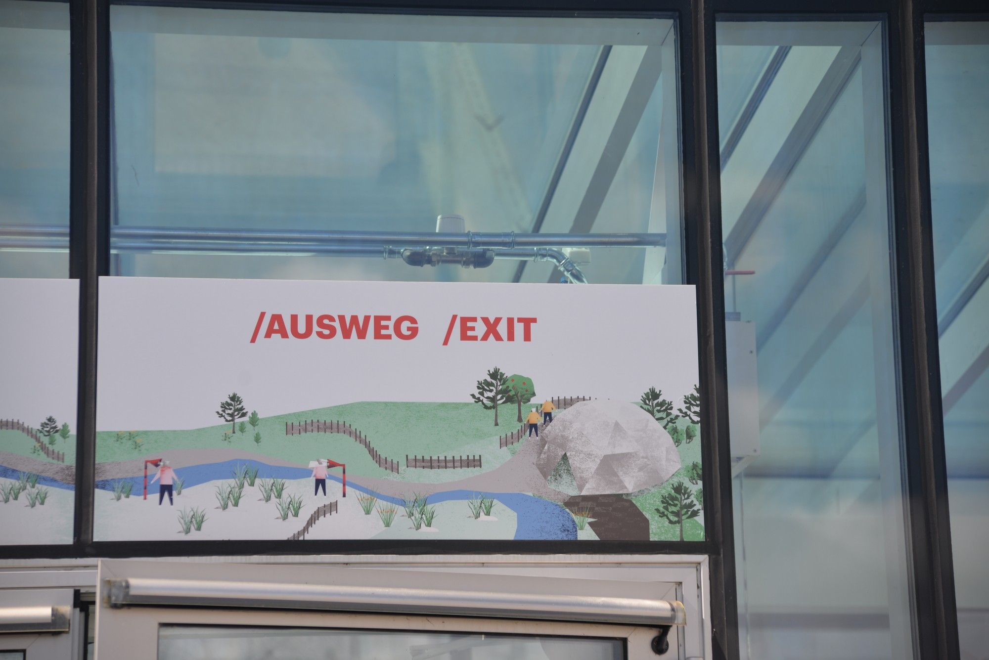 Ein Schild an einem Fenster. Der untere Teil stellt eine Landschaft dar, ein Fluss schlängelt sich durch, ein Grashang mit ein paar einzelnen Bäumen und vereinzelten Menschen.
Auf dem Schild steht in roten Großbuchstaben: AUSWEG / EXIT.