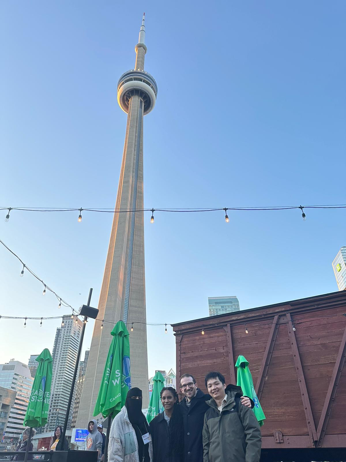 @raheemajavid.bsky.social, @hartwakeland.bsky.social, craig poskanzer, and @huangjiawen.bsky.social stand in front of
 the CN Tower