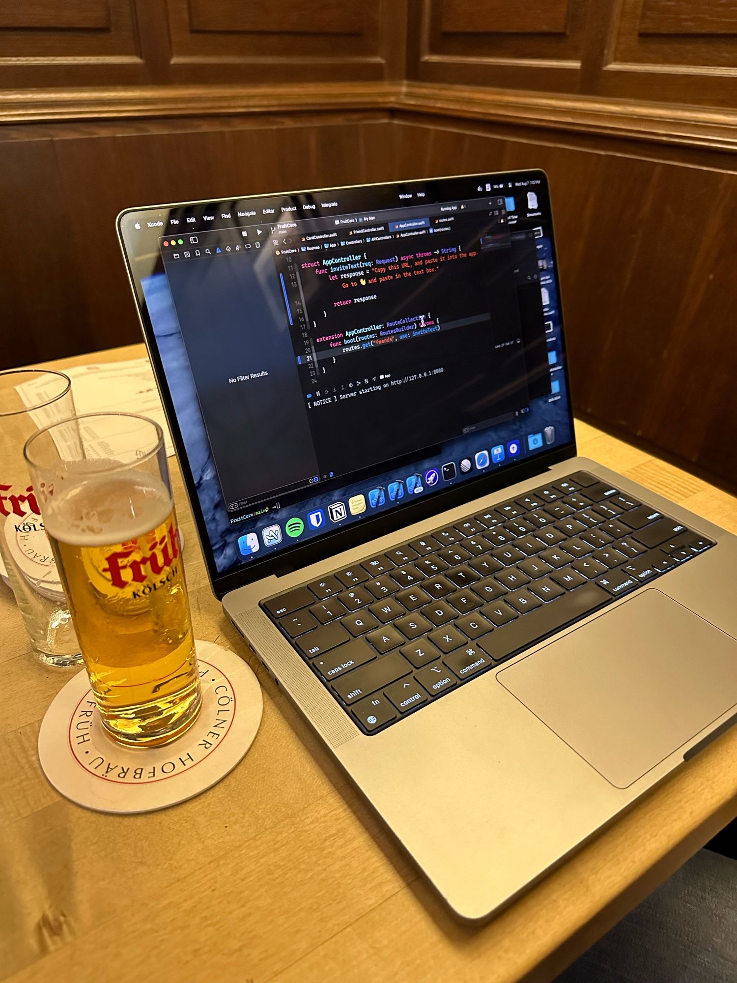 MacBook Pro with Xcode on the screen and an empty 0,2 L kölsch, and refilled one next to the MacBook