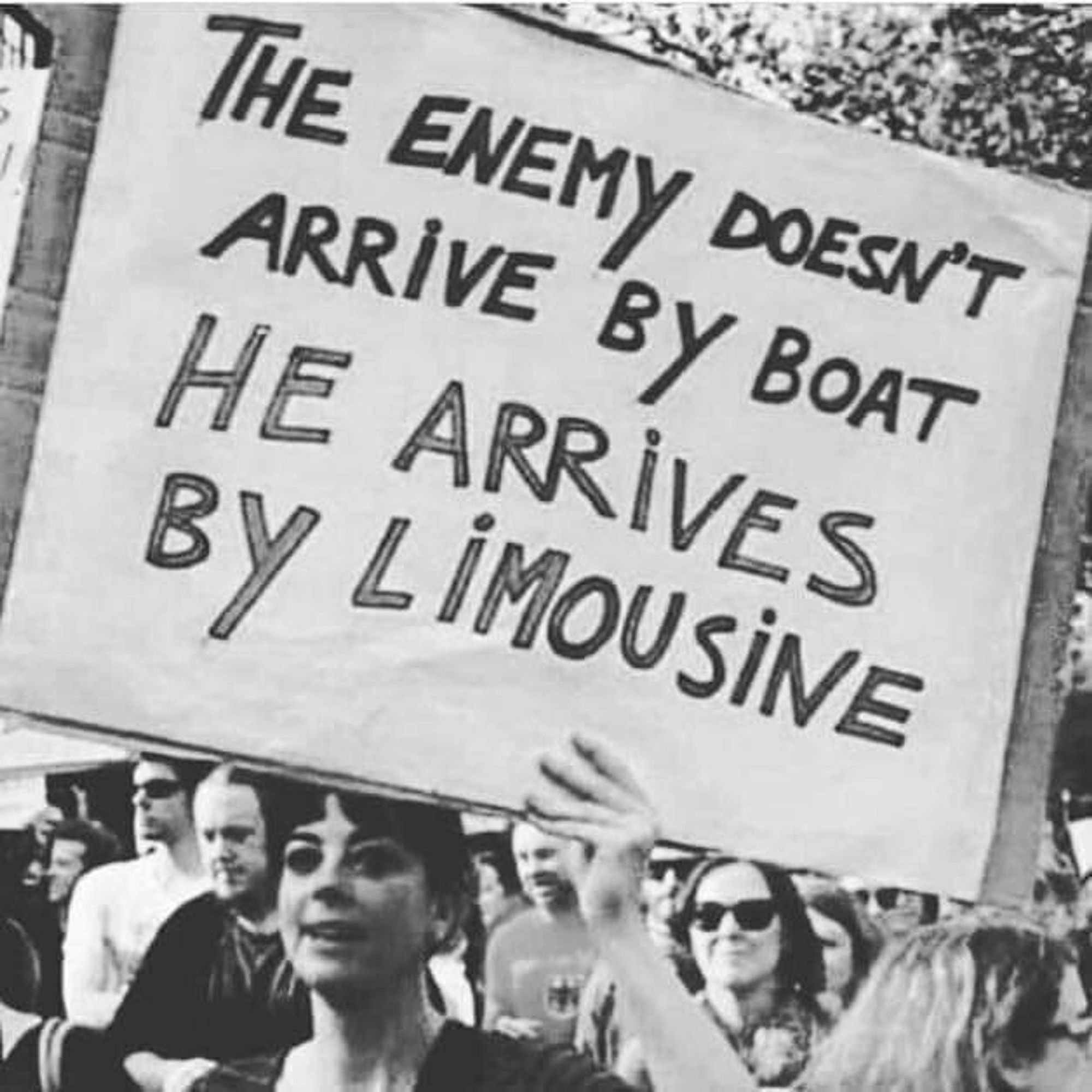 B/W-photo of a woman holding up a sign. The sign says „THE ENEMY DOESN'T ARRIVE BY BOAT HE ARRIVES BY LiMOUSiNE“