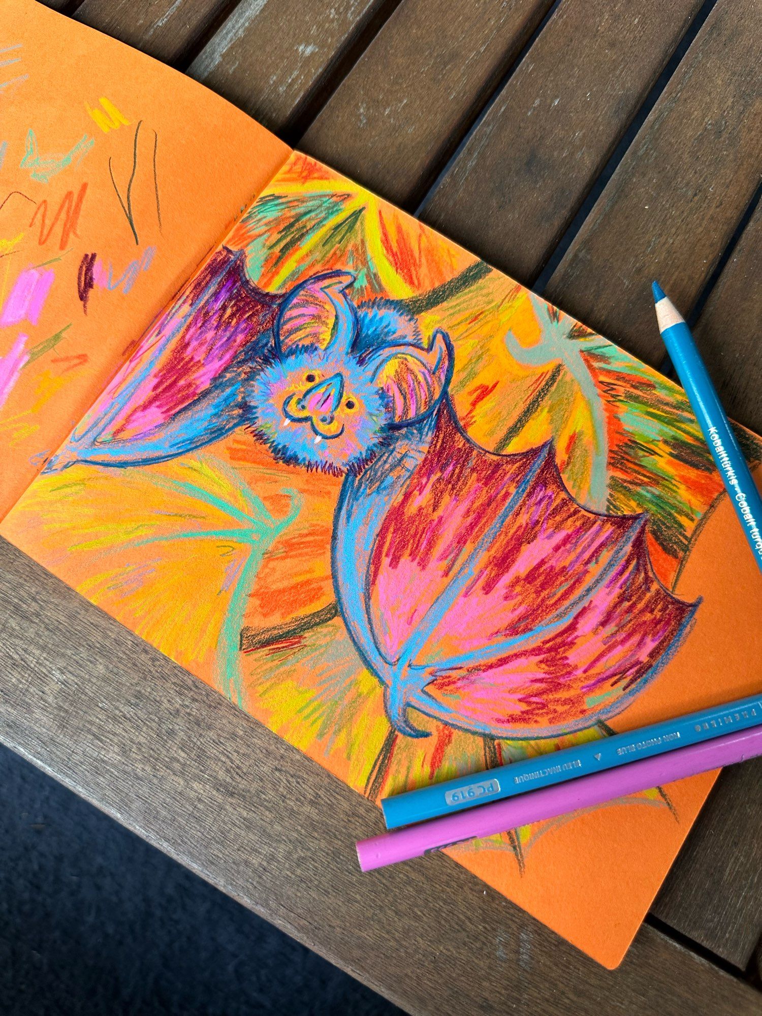 Photo of an open sketchbook with bright orange paper. The sketchbook is sitting on a wood coffee table. There’s a colored pencil drawing of a brightly colored blue bag with reddish-pink wings and ears and splashes of yellow on its ears and face. Two blue and one pink colored pencils lay on top of the paper, framing the image.