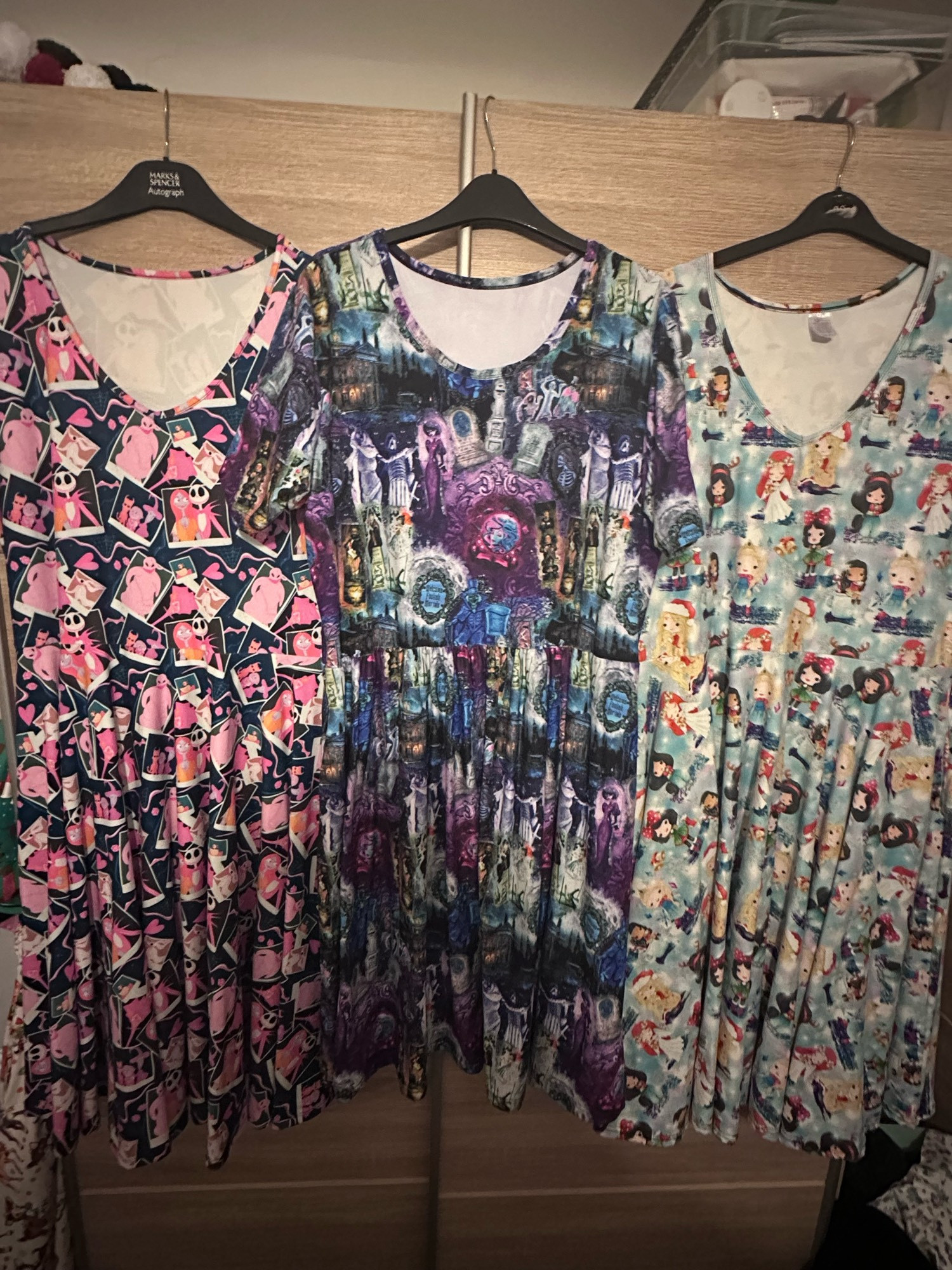 Three dresses hang from a wardrobe  door. One is pink and nightmare before Christmas themed, the next is Disney haunted mansion themed and the last is Disney princesses dressed in winter clothes print.