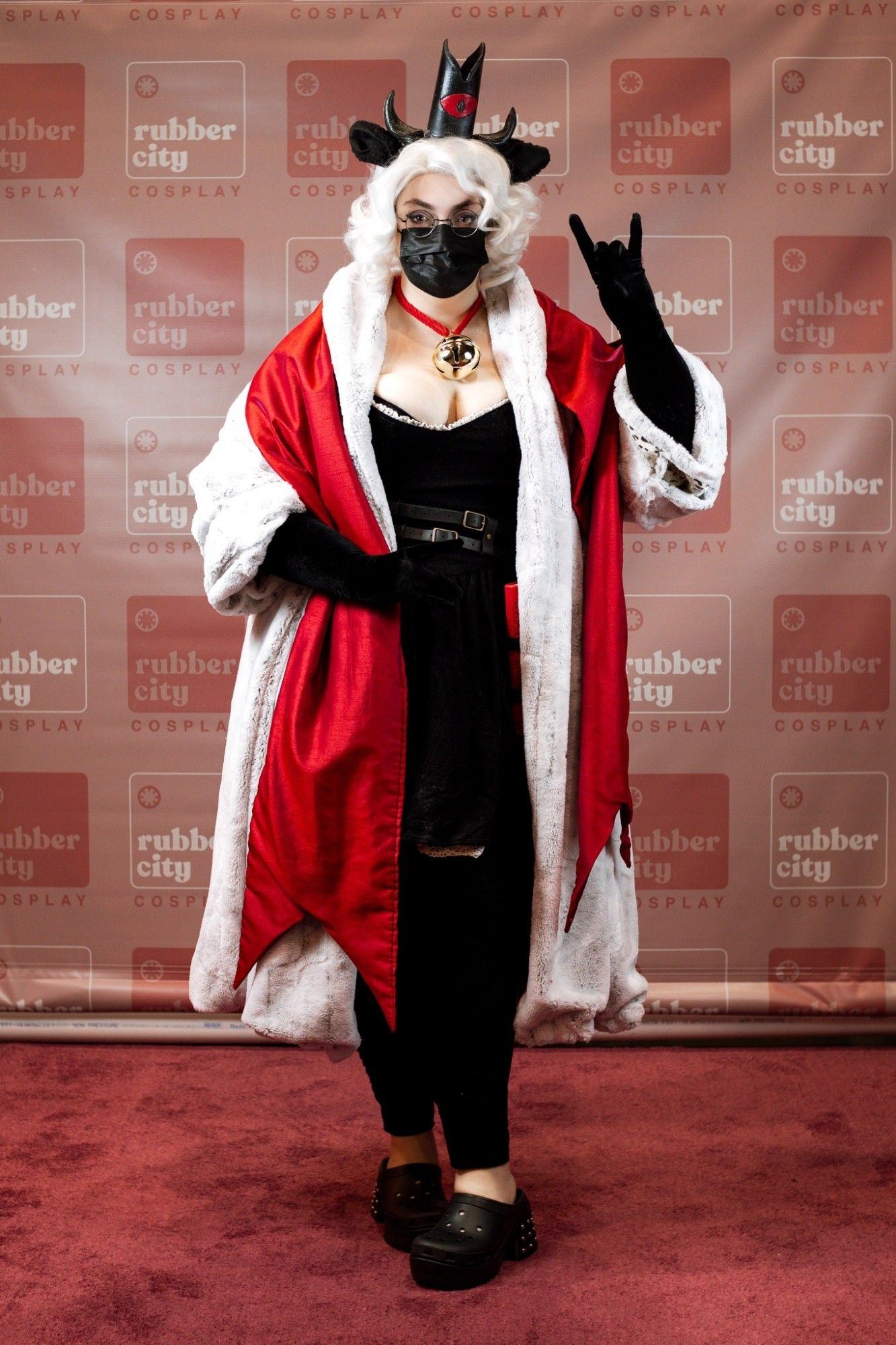 My 'cult of the lamb' cosplay, a white haired woman in black gloves and mask has a massive white fur coat and red vestments. A crown and sheep ears sit on her head