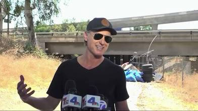 A photo of Gavin Newsom at the press conference dressed in a tight black v cut t-shirt, black aviators, and a black cap, smiling as he answers questions