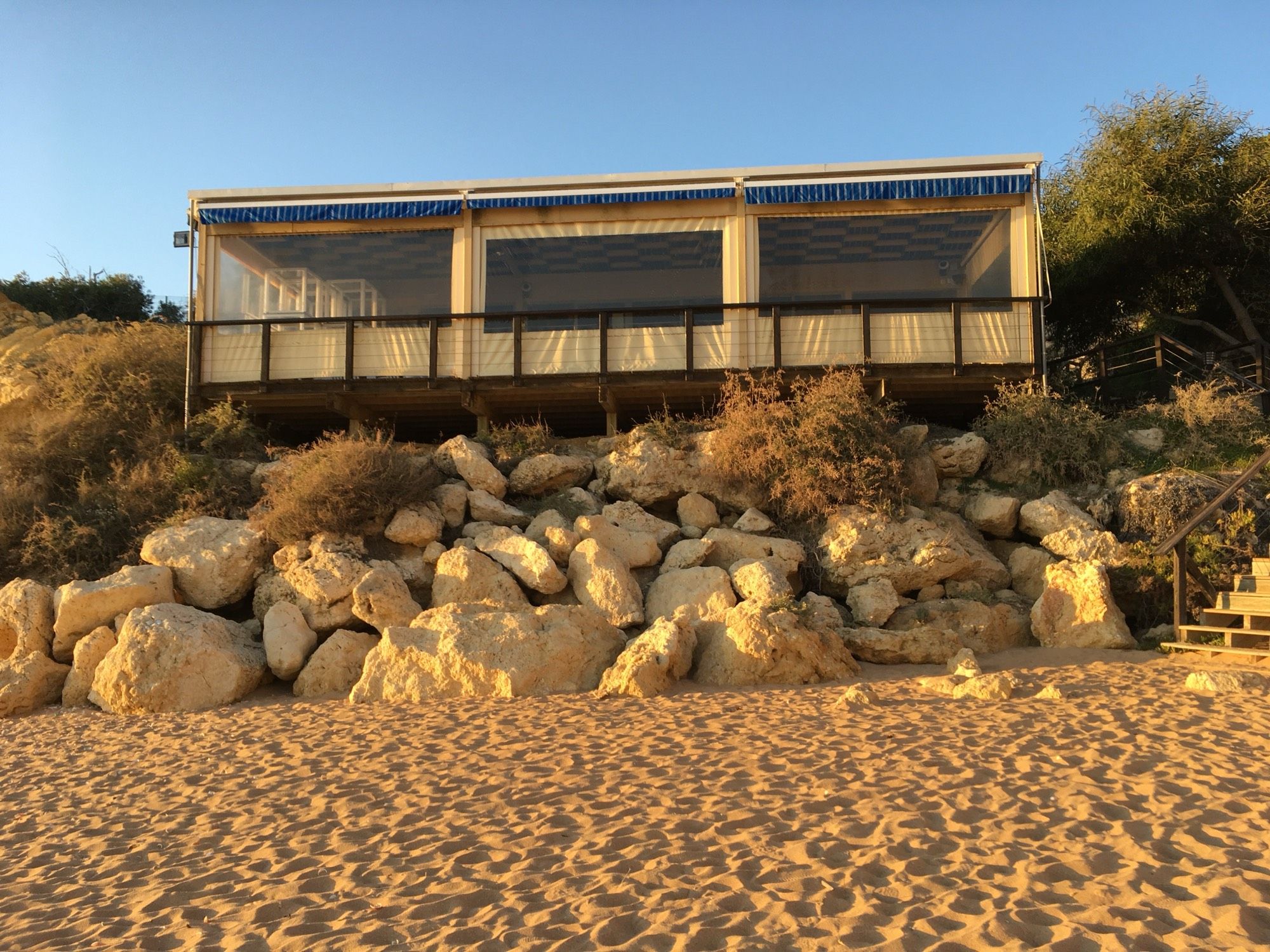 Strandbar auf Felsbrocken, davor Sand in der Abendsonne.