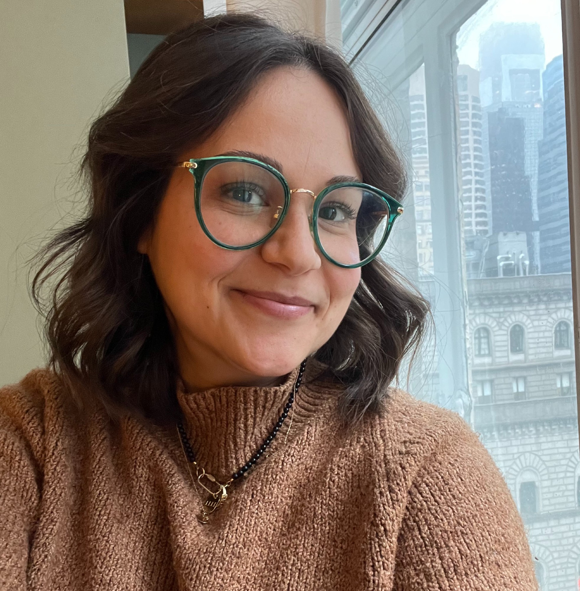 a photo of jamie wearing green glasses, a brown sweater, and black beaded necklace with a pendant hangs around her neck, with a city skyline visible through a window.