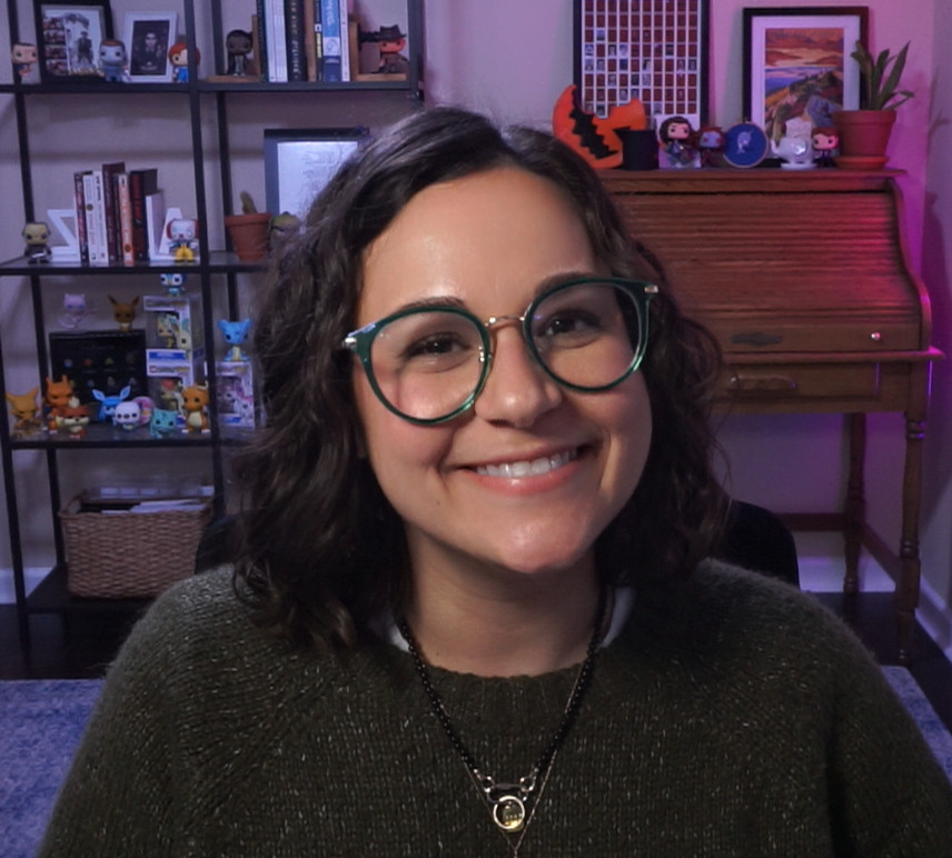 a photo of jamie with shoulder-length dark hair and glasses, smiling warmly at the camera. she is wearing a textured green sweater and a necklace with a circular pendant. In the background, a cozy room is visible, including a bookshelf filled with books, framed pictures, plants, and various figurines. There is also a wooden roll-top desk with additional decorations on top, including small plants and photographs. 