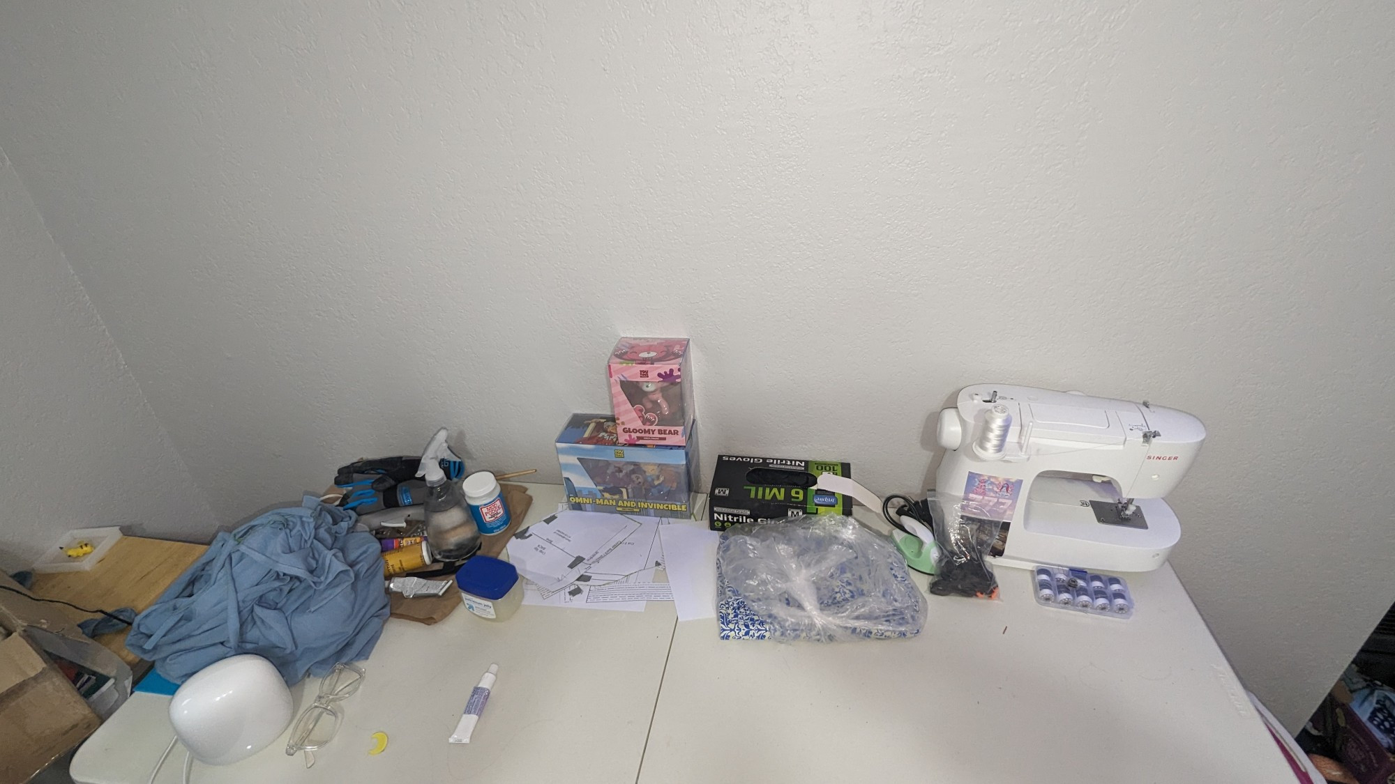 up close of my craft desk; theres nitrile gloves, a sewing machine,, mod podge, various adhesives, fabric, and a fabric pattern on the desk. 