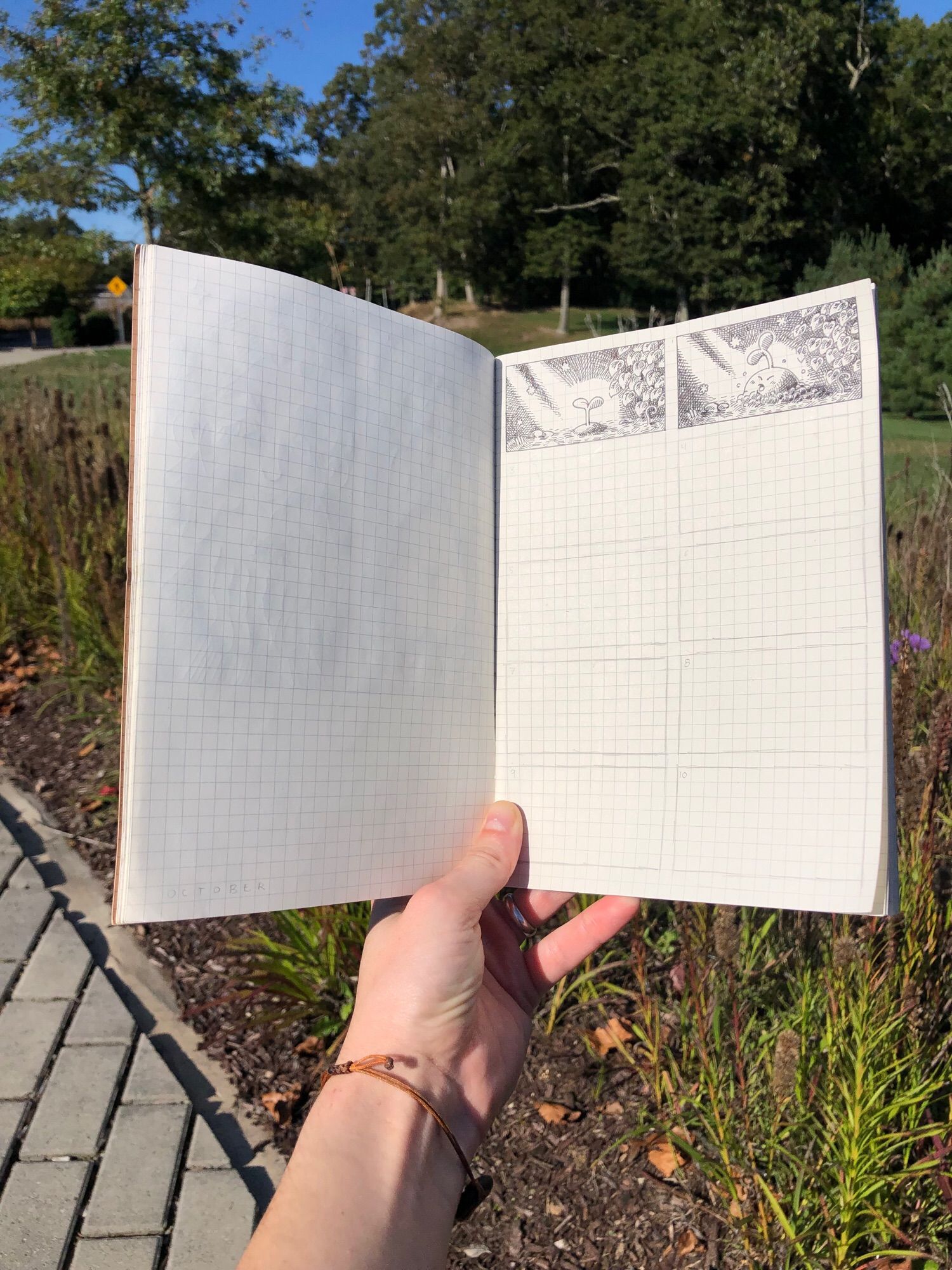 Photo of a hand holding open a sketchbook at a park. There are 2 out of 10 comic panels that have been drawn in.