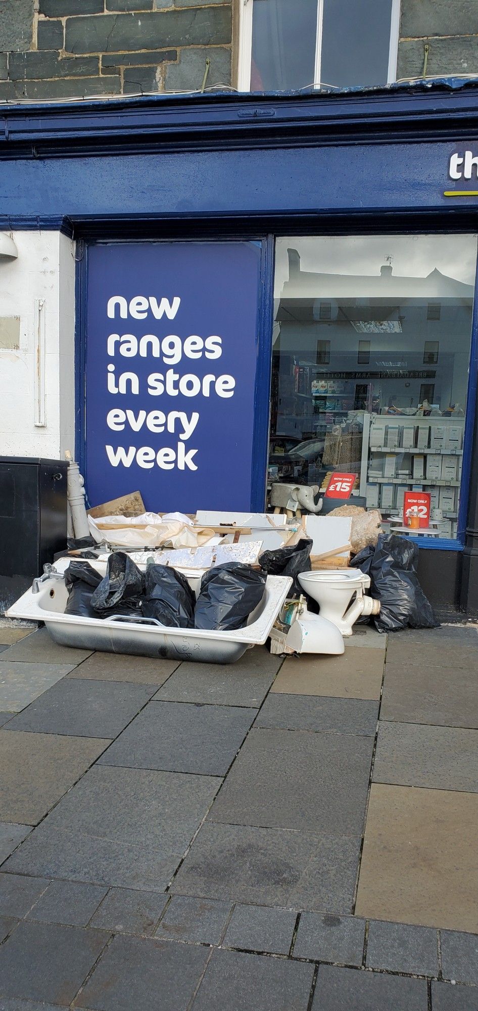 Discarded bathroom suite and bags of rubbish in front of a store sign reading "New ranges in store every week".