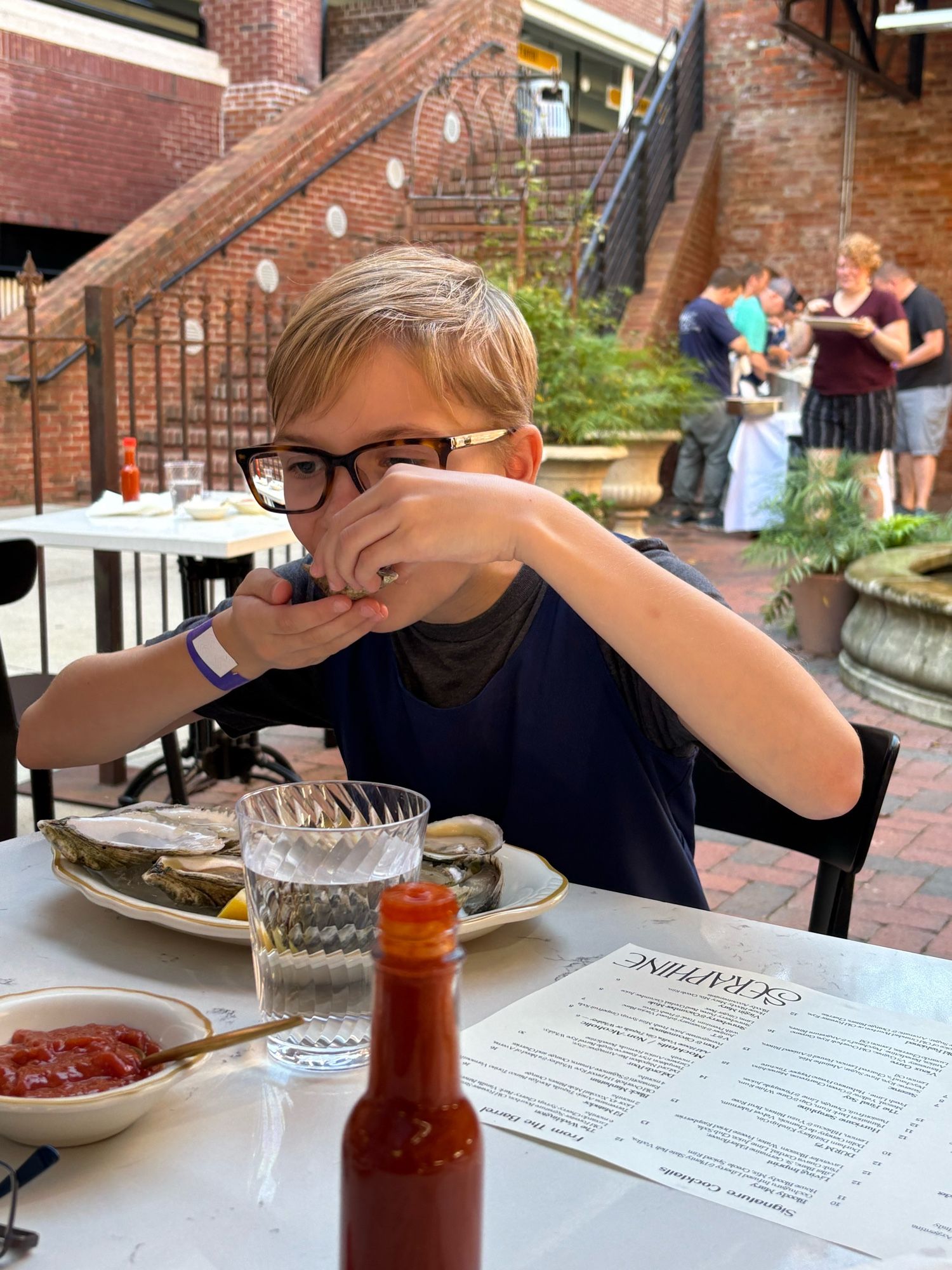 A child with short blond hair and glasses sits at a table in a brick courtyard, tipping a raw oyster on the half shell into her mouth. She’s eating them as fast as she can to make sure she gets her “fair share”. She later hit her parents up for some of their oysters.