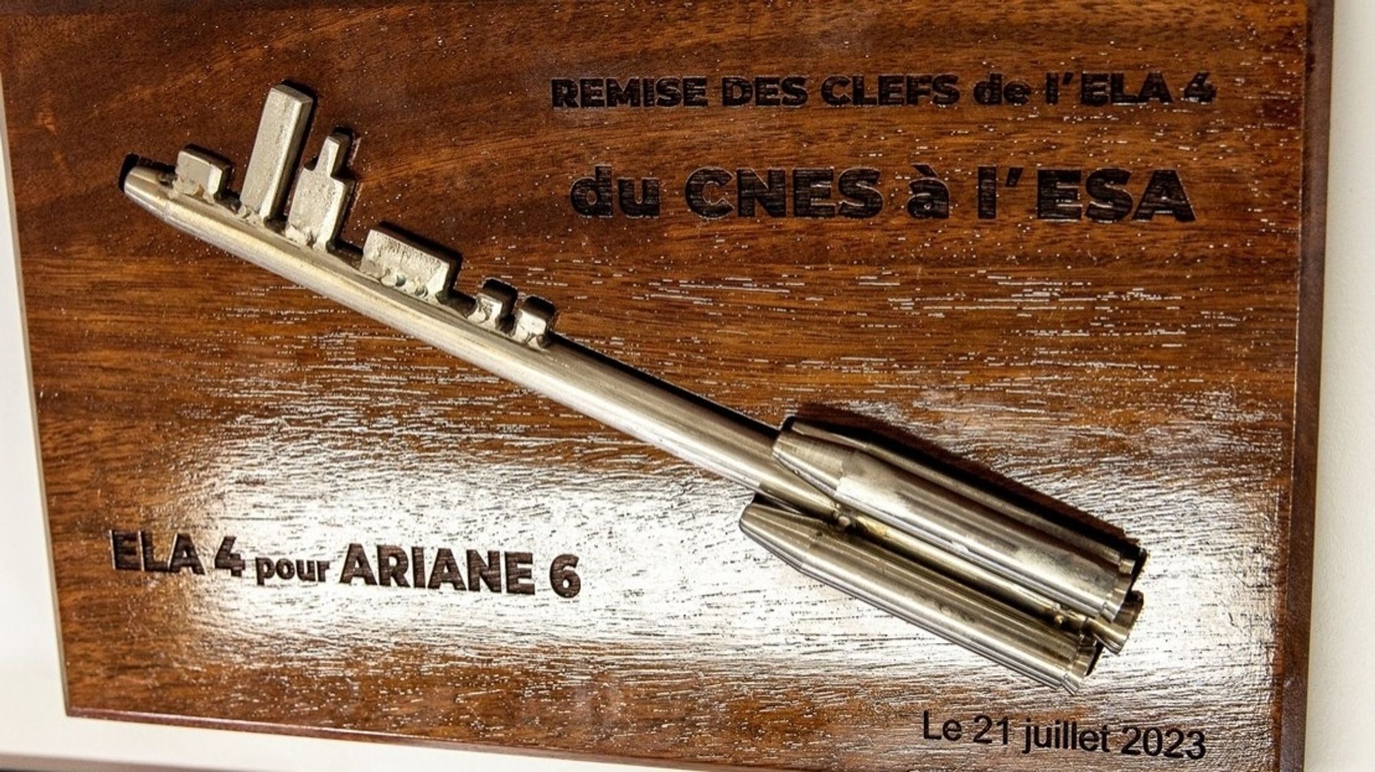 Sur une plaque de bois sombre est fixée une clef symbolique : la base de la clef comporte 2 boosters et les dents de la clef représentent les bâtiments d'ELA 4.
Sur la plaque figure (de haut en bas, de gauche à droite) :
- Remise des clefs de l'ELA 4 du CNES à l'ESA
- ELA 4 pour ARIANE 6
- Le 21 juillet 2023