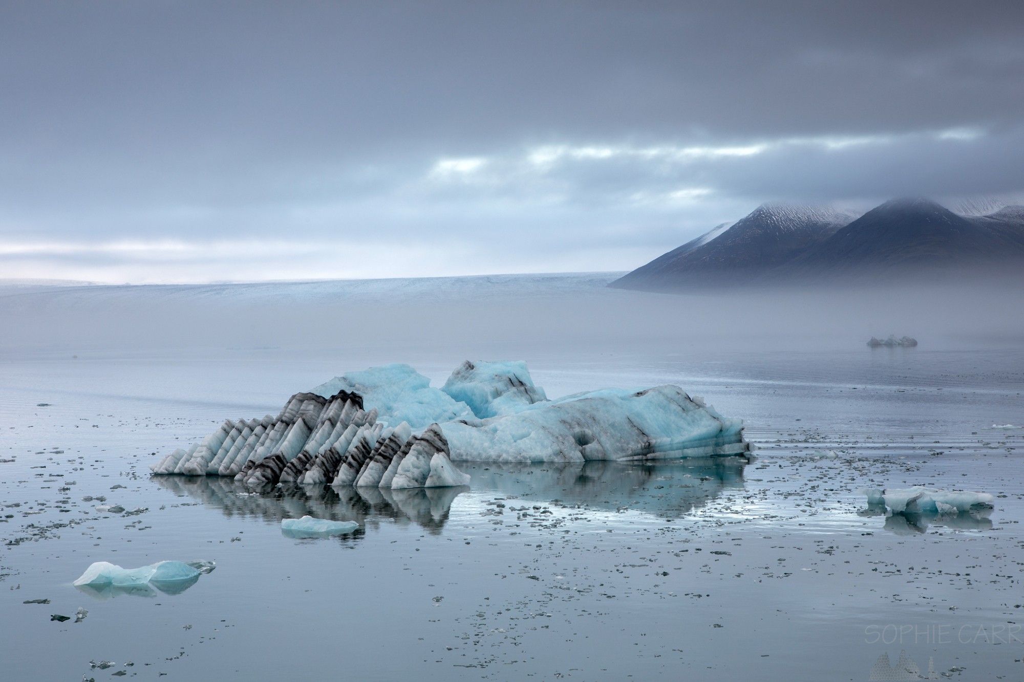 A couple of interesting icebergs sit in the Jökulsárlón glacial lagoon. The water is very still and behind there is a layer of fog, with some pointy mountains behind.