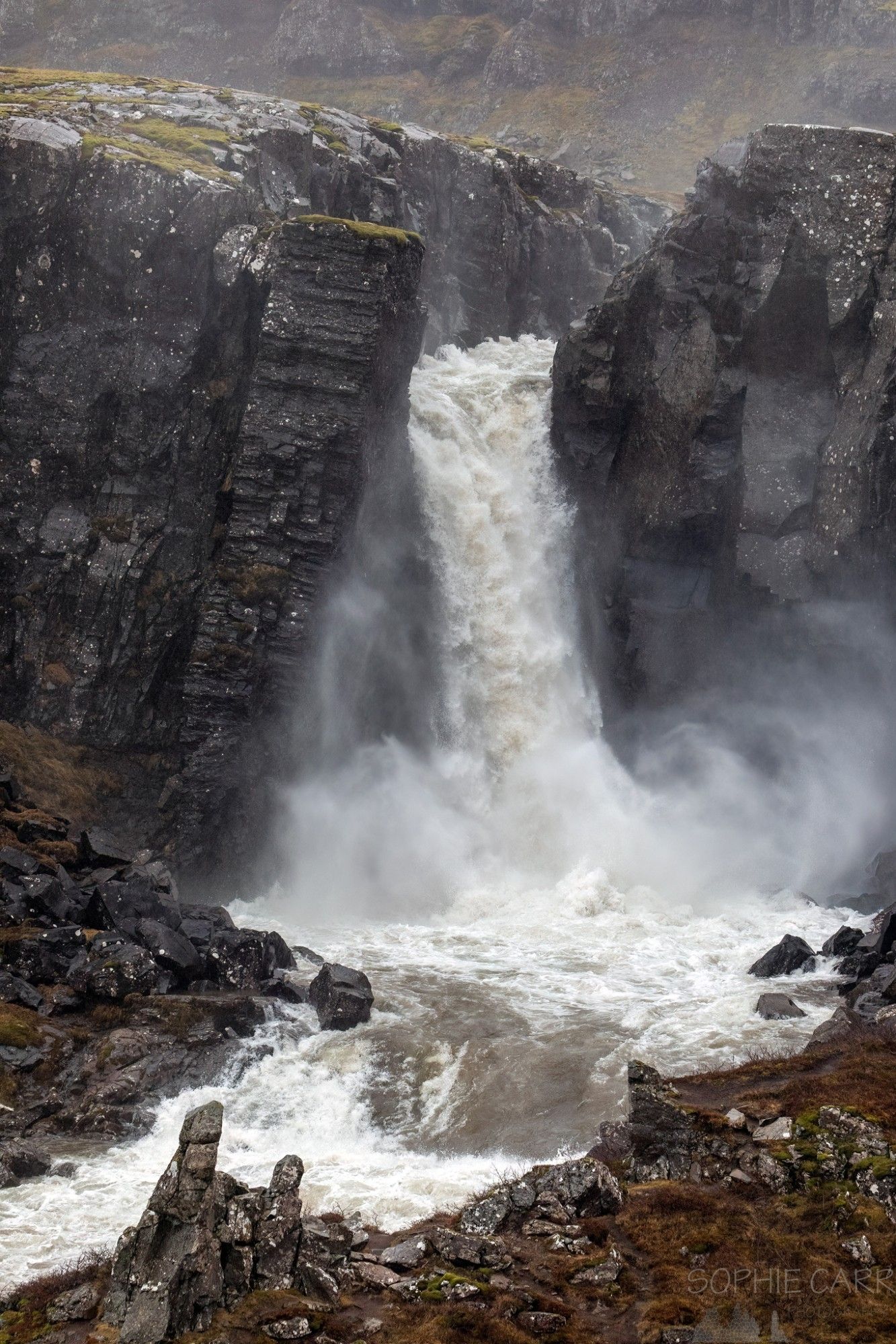 A raging waterfall comes through a gorge with big black rocks; spray from the waterfall is rising from the bottom of the falls. Some brown grasses in the foreground.