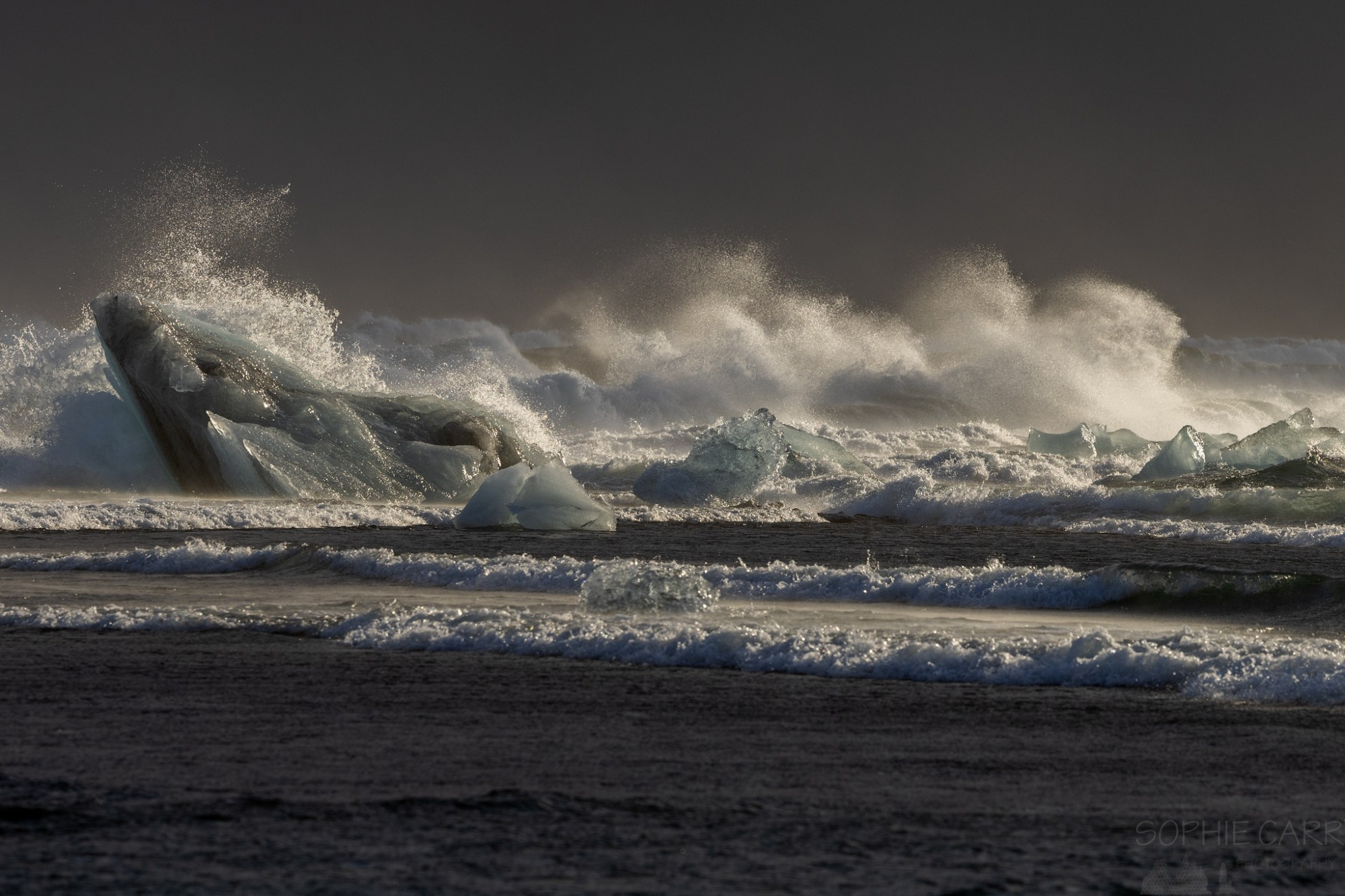 Waves crash over icebergs and there's lots of spray in the air. There is sun on the ice and the crashing waves, but the sky is dark behind.