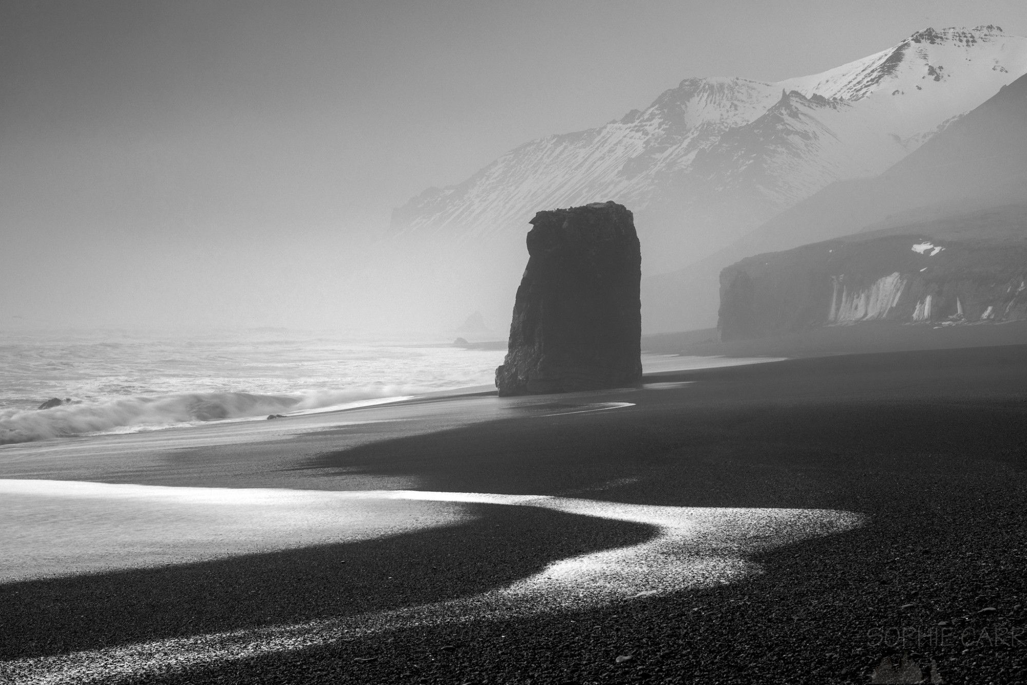 A black sand beach has some white froth from a receding wave, and a large stack rises on the beach. There are some icicles on the small cliff behind, and in the distance are some snowy mountains, just visible through a light fog. Photo converted to black and white.