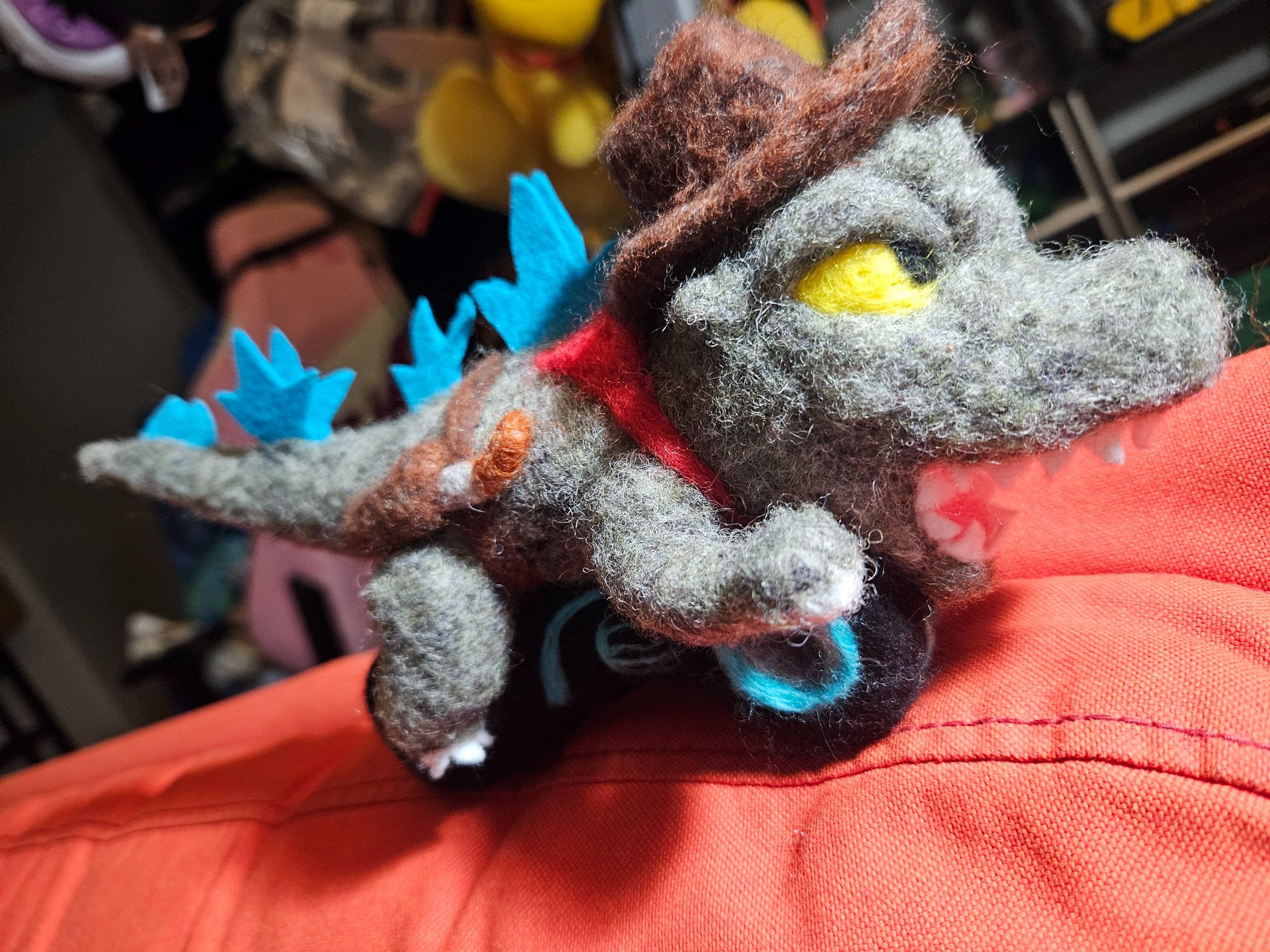 A needle felted sculpture of a chibi Godzilla on a lightcycle from Tron Legacy, wearing a cowboy hat, gun holster, and red bandana. 