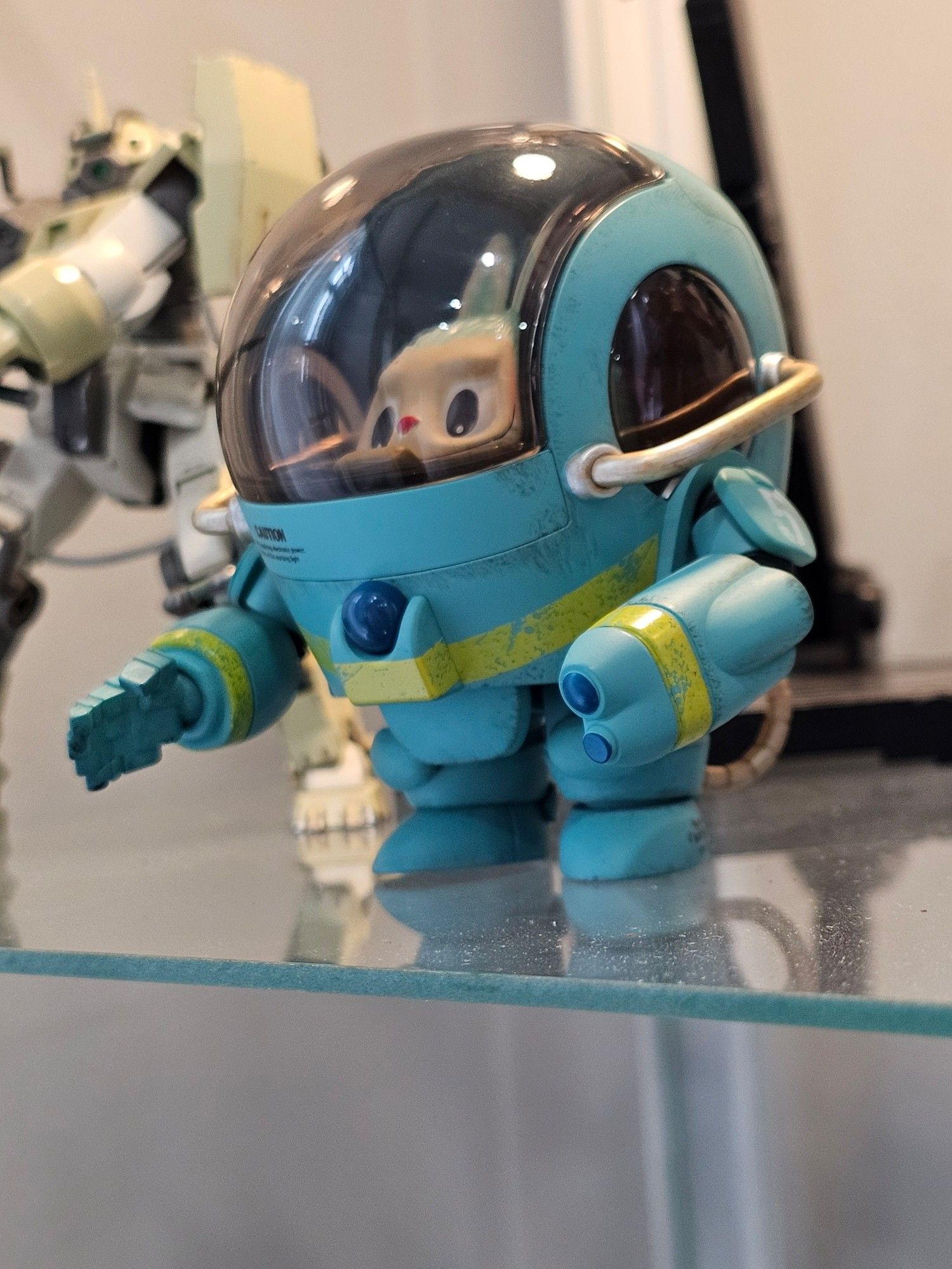 Figurine of a small teal blue slightly-beat-up mech with a yellow stripe across the middle, and a big glass bubble cockpit canopy through which is a distorted view of a cute little creature. Photo.