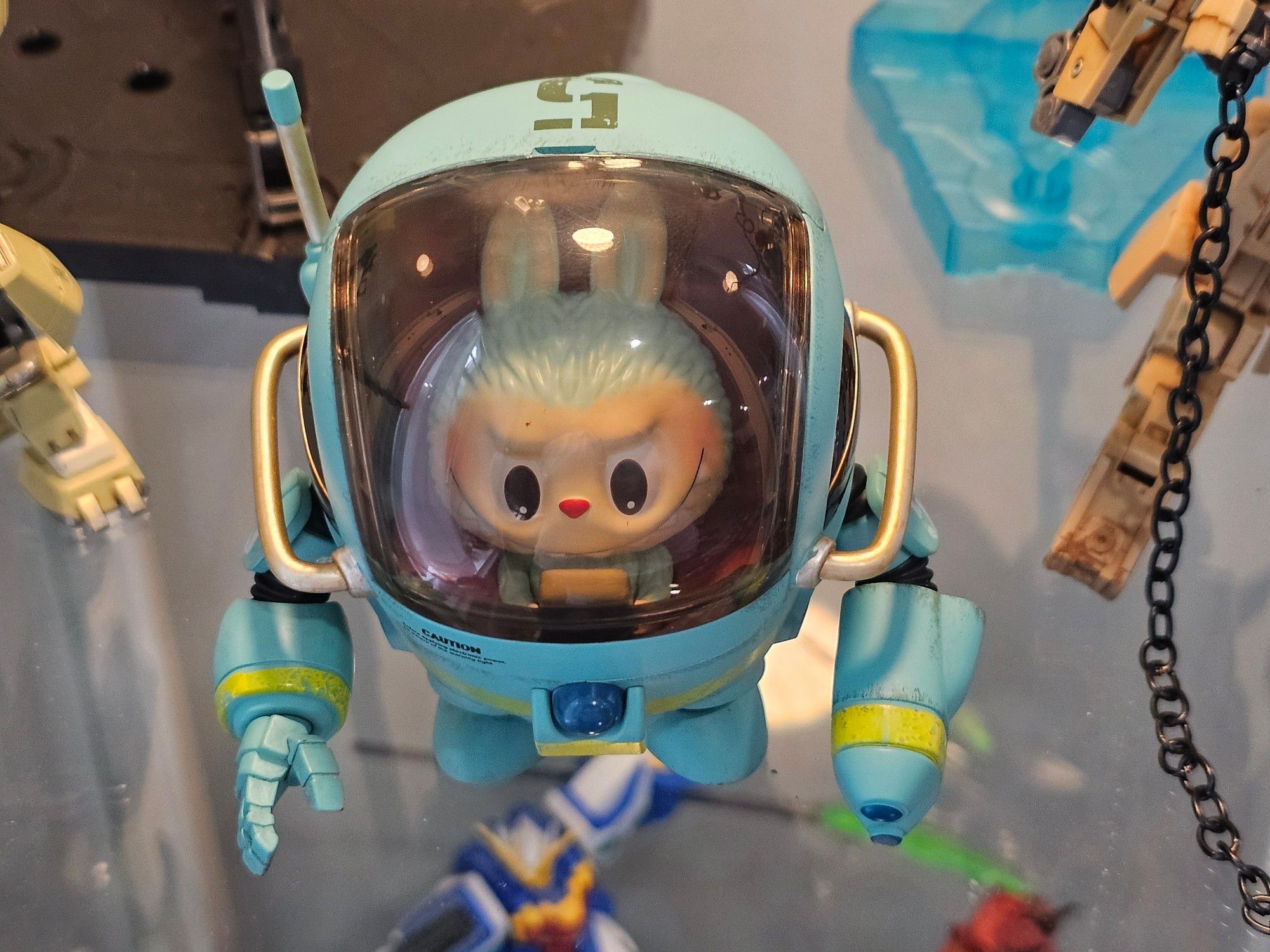 View of the mech figurine from above. The figure of the critter inside, named Ququ, is like a baby in a fuzzy teal blue bunny costume with lots of sharp teeth. They're playing a Gameboy with a look of excited concentration. The mech has one humanoid hand and one double barrelled gun hand. Photo.