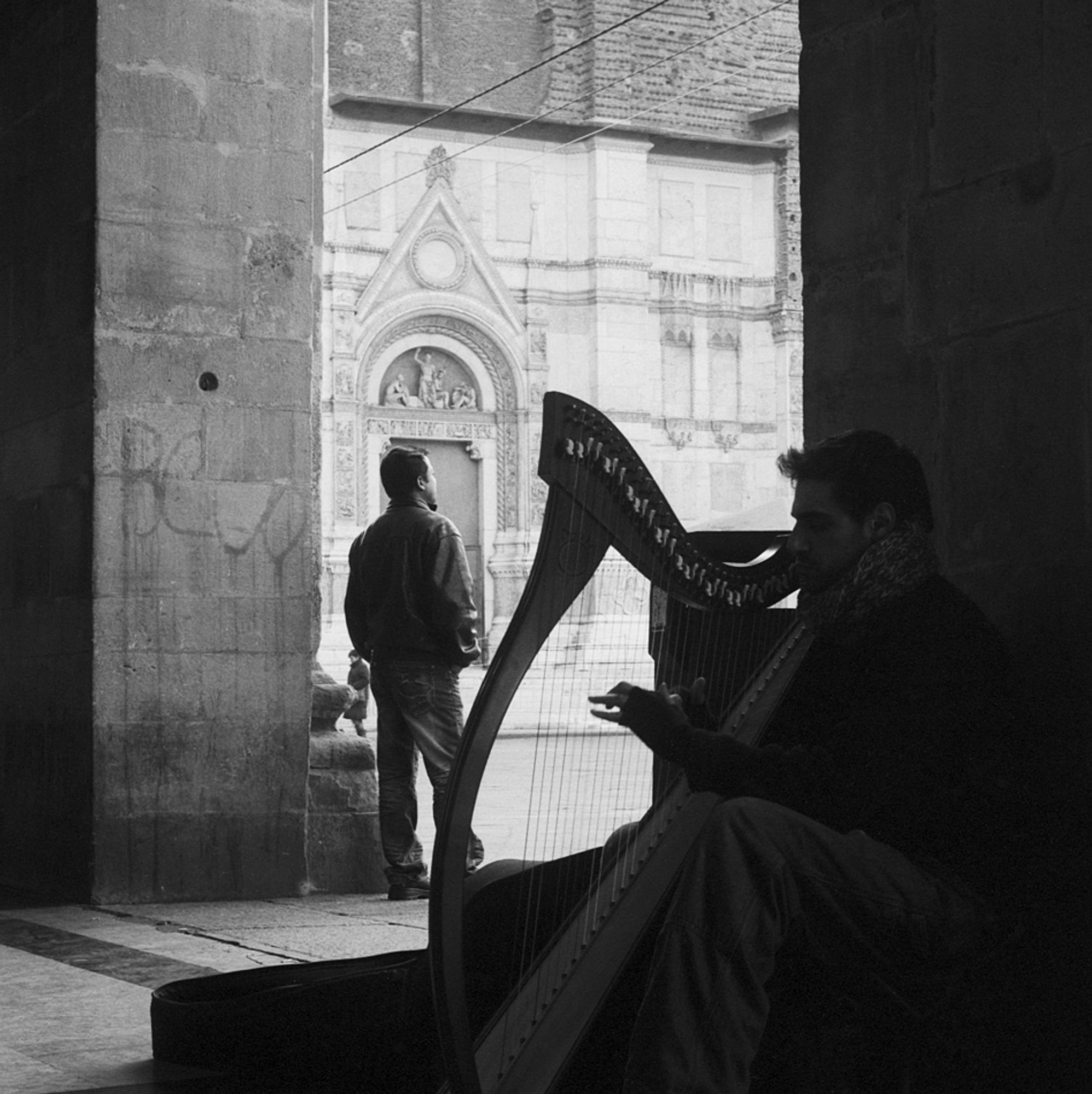 Harpist busking on the day before Christmas Eve under the portico of Via dell'Archiginnasio in Piazza Maggiore with the Basilica di San Petronio in the background while a man stands listening, Bologna, Italy, 2008.