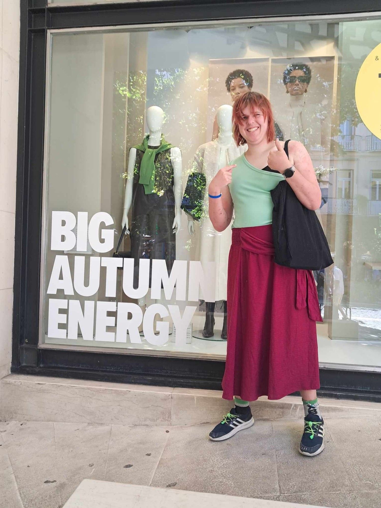 Autumn standing by a Marks and Spencers window display, next to the words "Big Autumn Energy". 
She is wearing blue Adidas trainers with green elastic laces,a green one shoulder crop and is carrying a black Primark Pride bag