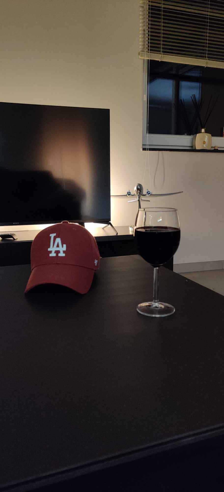 Un verre de vin sur une table de salon avec une casquette de Baseball bordeaux taguée LA...