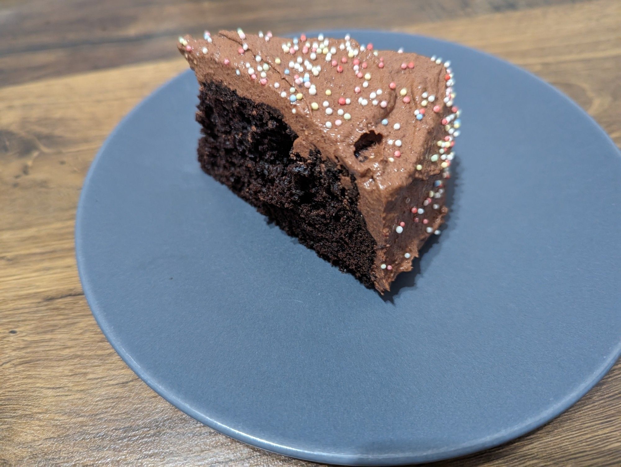 Auf einem dunkelblauen Teller steht ein Stück Schokokuchen. Dabei sieht man den dunklen Teig, welcher mit Schokosahne überzogen ist. Auf der Schokosahne sind bunte Streusel.