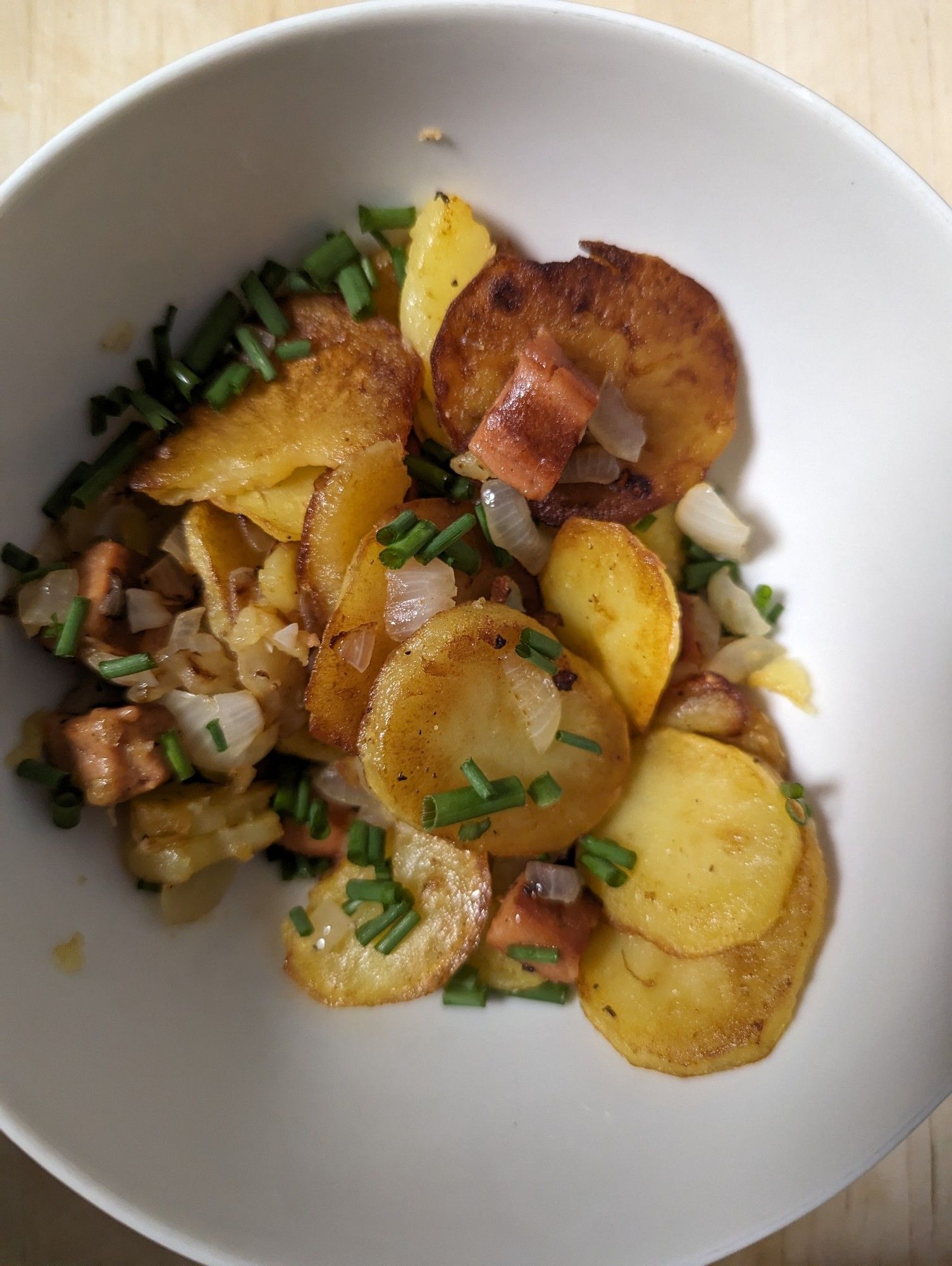 In einer weißen Schüssel sind die fertigen Bratkartoffeln mit Leberkäse und Zwiebeln zu sehen. Als Topping ist geschnittener Schnittlauch drübergestreut.