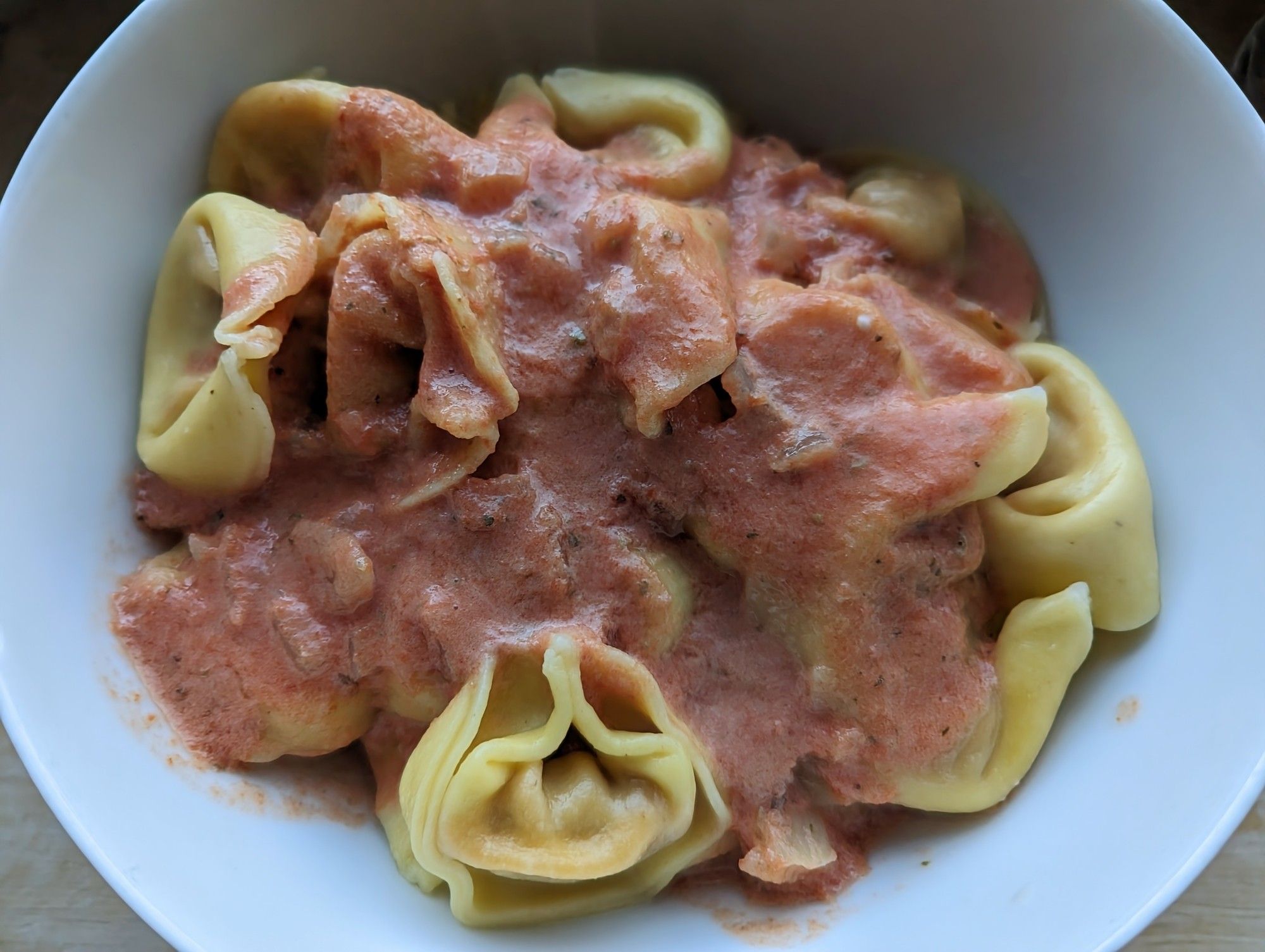 Weiße Schüssel mit gelben Tortellini gefüllt. Darüber rote Tomatensoße mit Zwiebelstückchen.