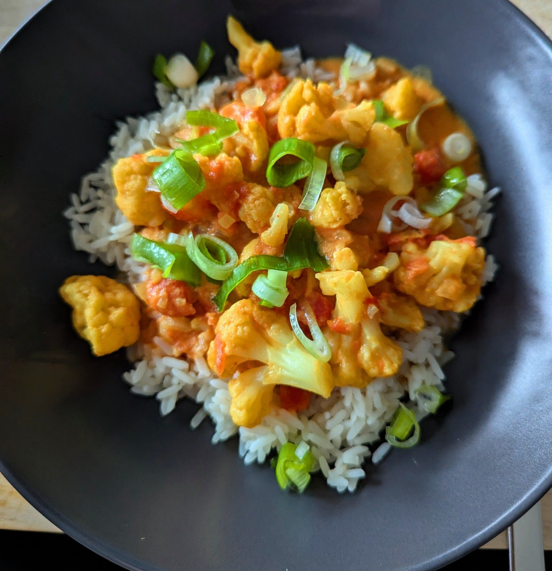 Das Bild zeigt eine Schüssel mit einem Gericht, das aus Reis und Gemüse besteht. Der Reis bildet die Basis, darauf liegt eine bunte Mischung aus Blumenkohlröschen, Tomatenstückchen und Frühlingszwiebeln in einer gelben Currysoße.